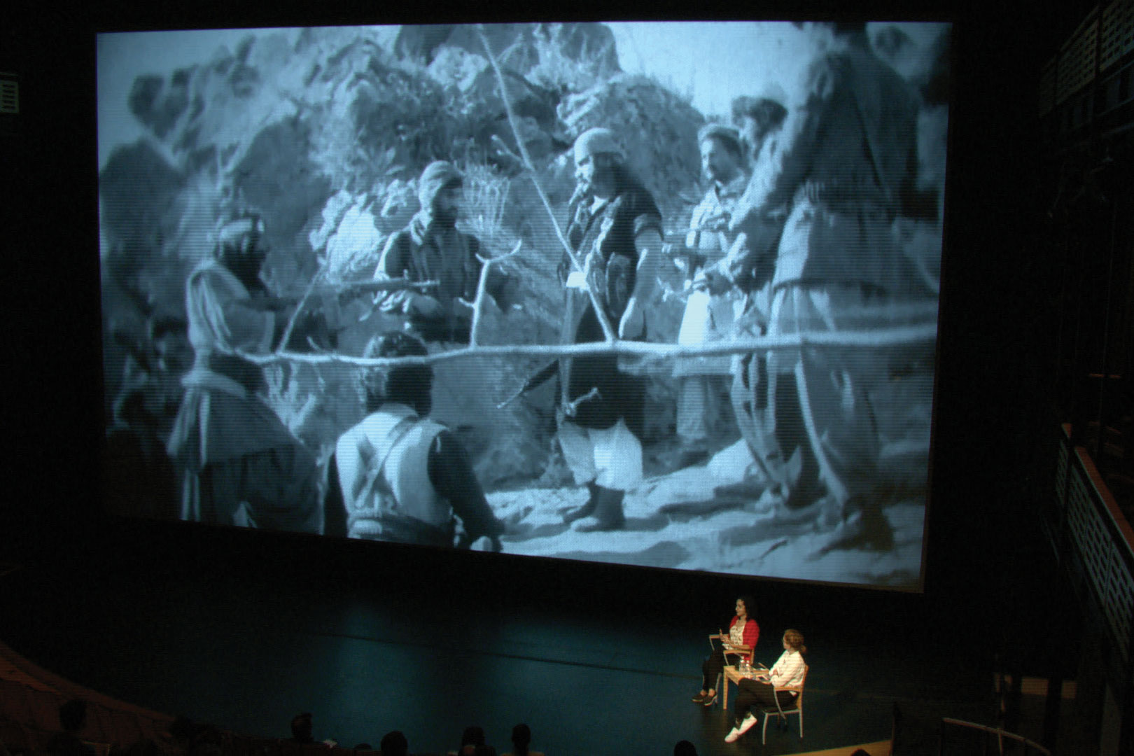 miriam ghani and Vic Brooks on stage in front of a large projection of an Afghan film. 