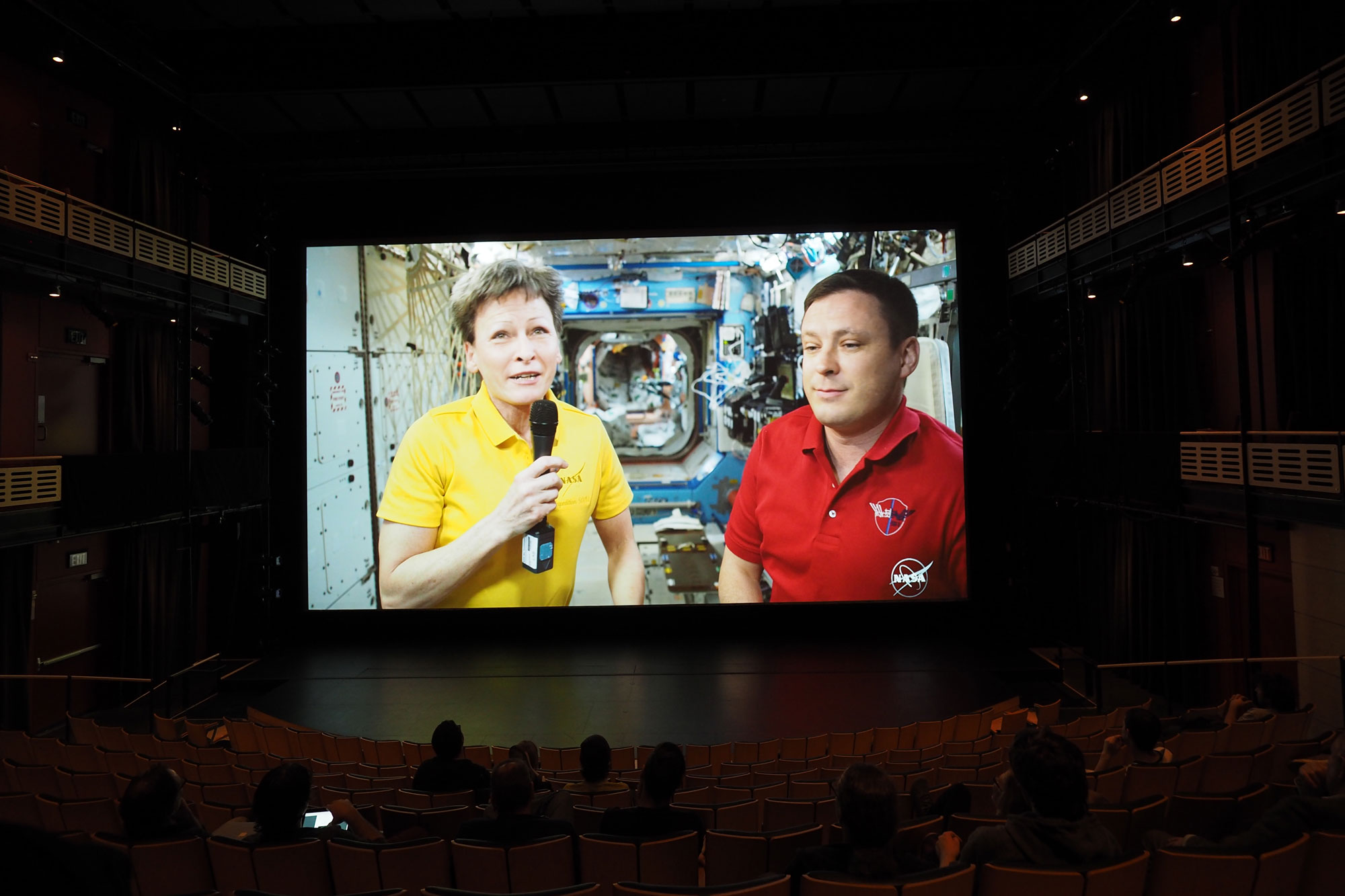 A female astronaut in a yellow polo holding a microphone and a male astronaut in a red polo talking via livestream to a silhouetted crowd.  
