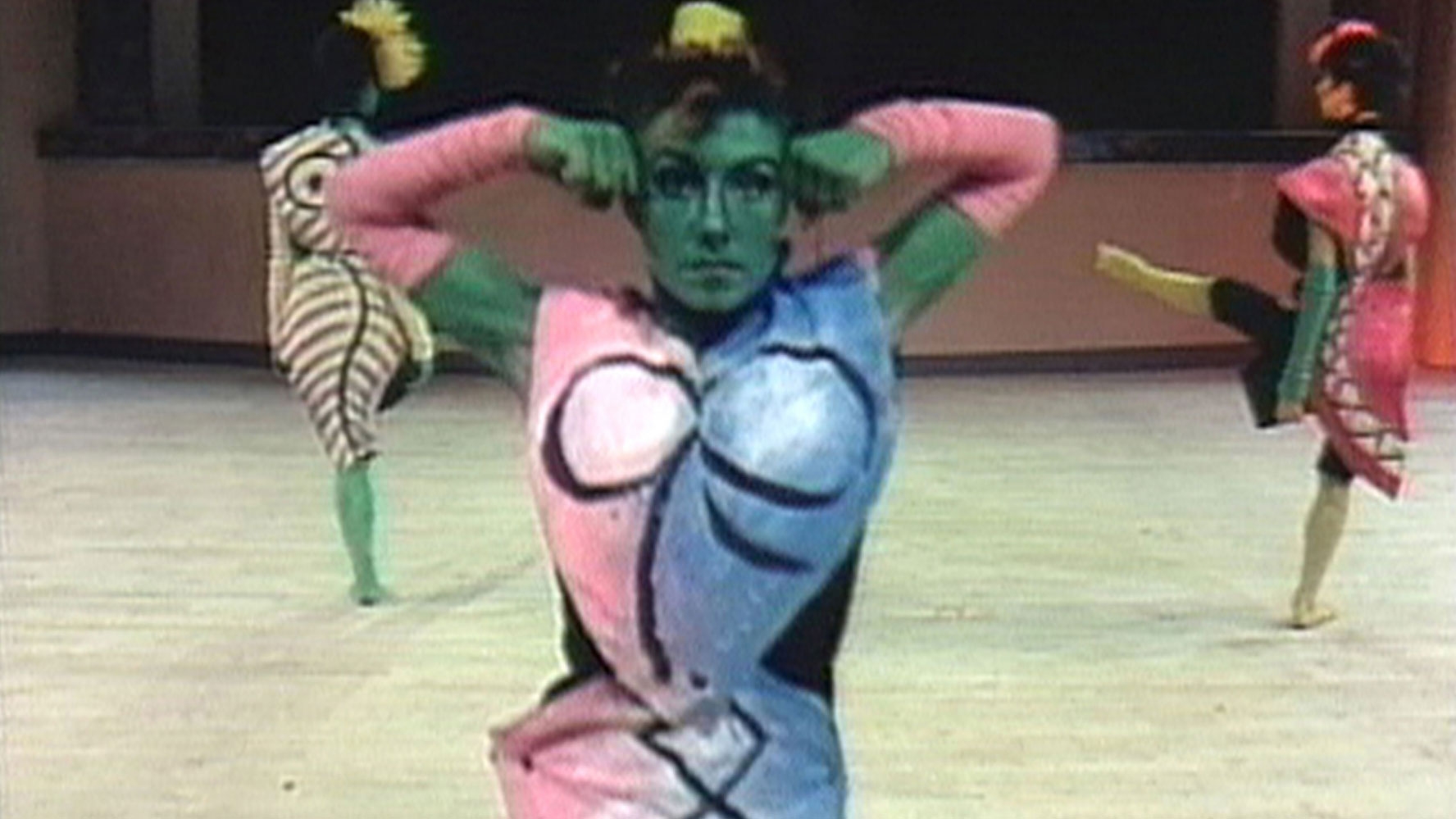 A woman painted green wearing a half pink, half blue costume with a Picasso inspired nude female form painted on her outfit. Two other female performers are in the background similarity dressed.  