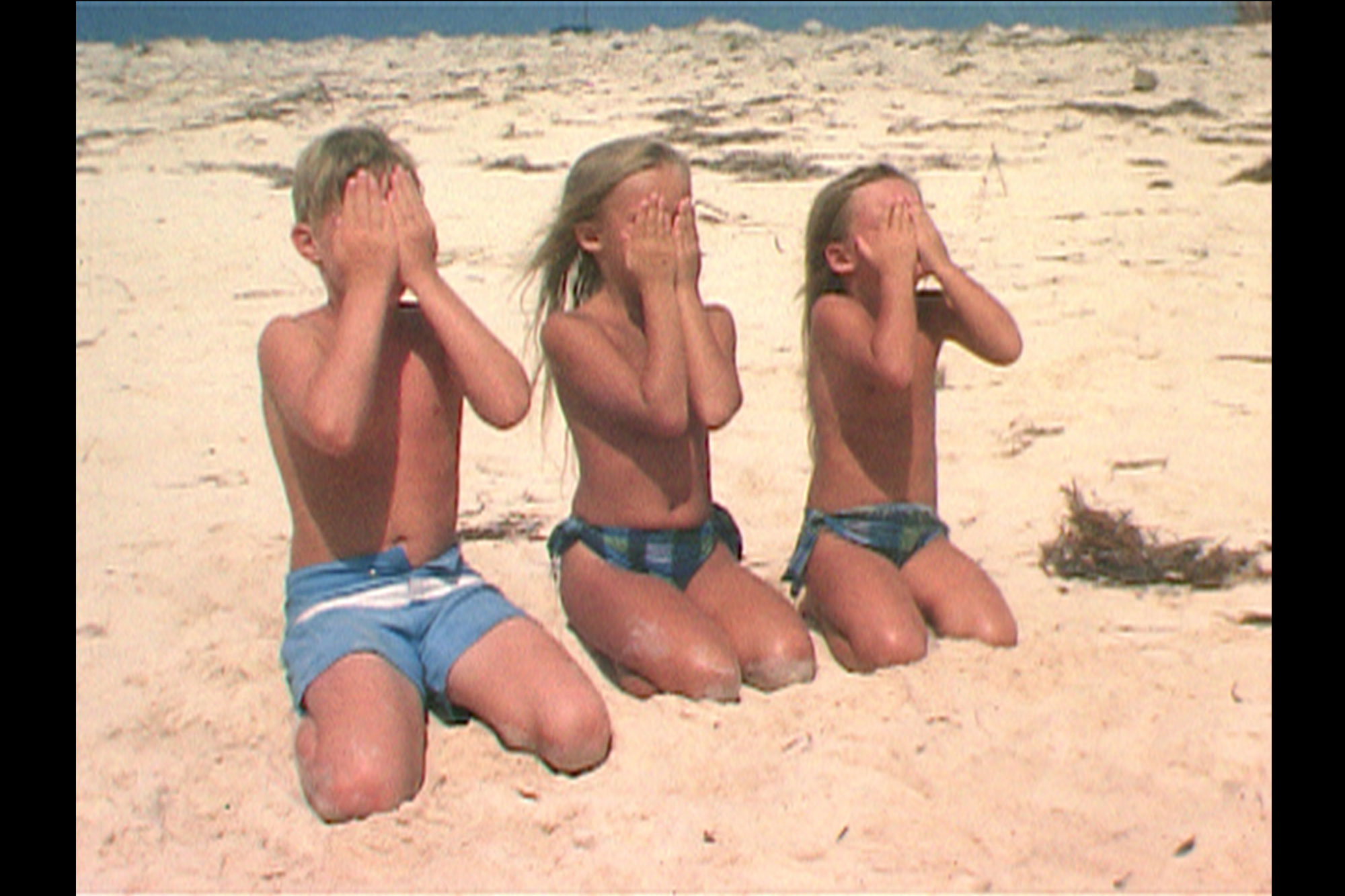 three blonde childern kneeling in the sand with their hands over their faces. 