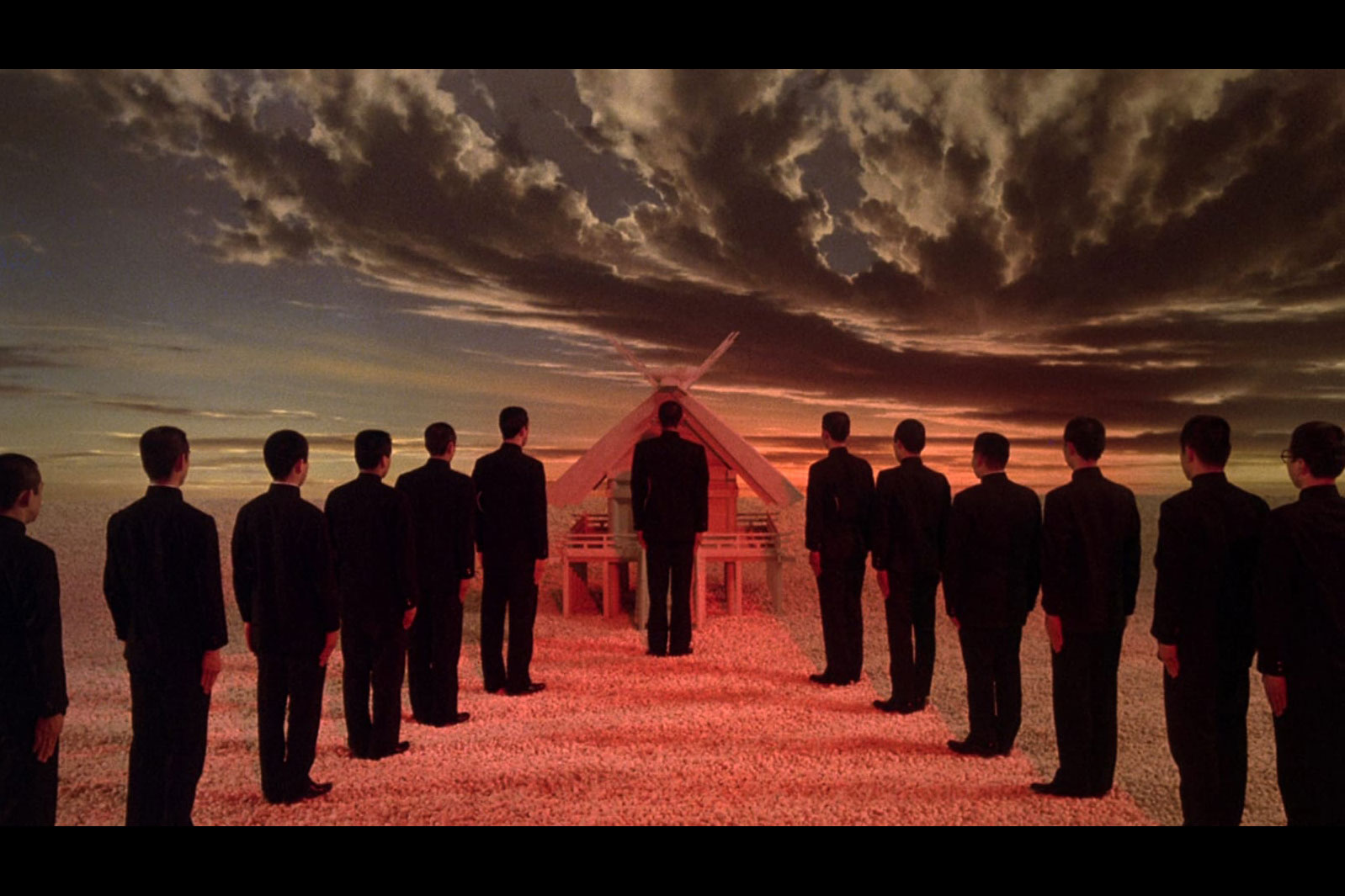 Thirteen men dressed in black in a surreal landscape standing with backs to the camera in a 'V' formation with one man at the point, facing an alter. 