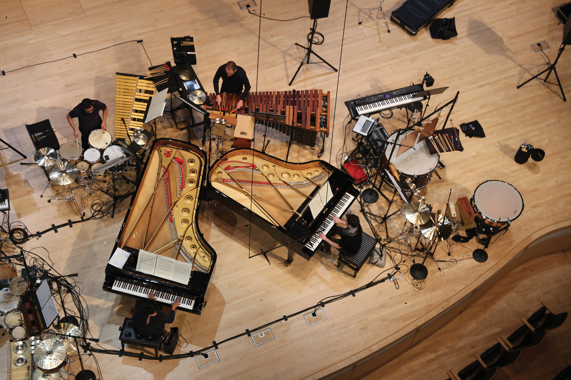 An aerial view of two people playing pianos and two people playing various percussion instruments on a clutter concert hall stage 