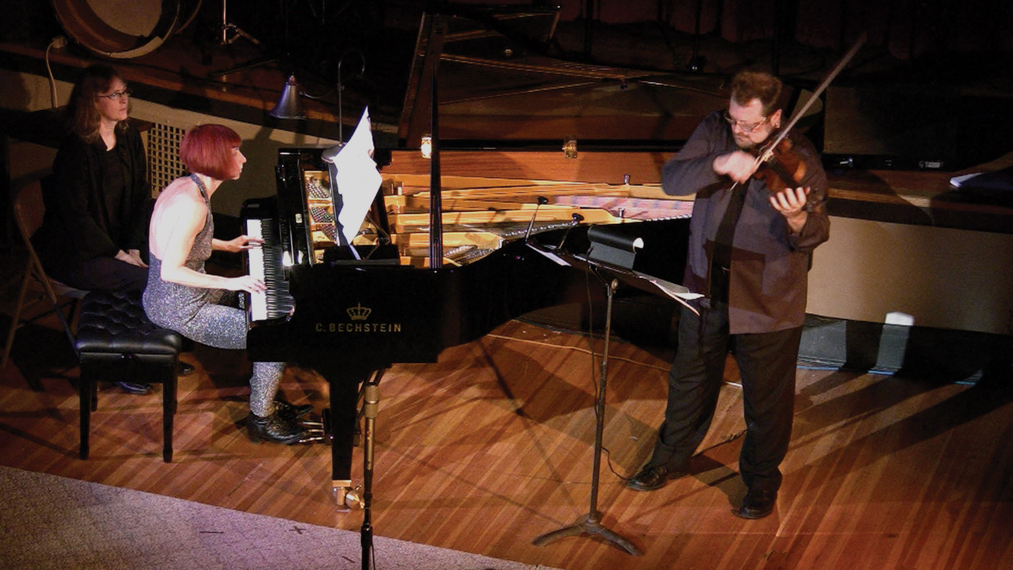A woman with red hair playing a grand piano on a wood stage as a man plays violin. 