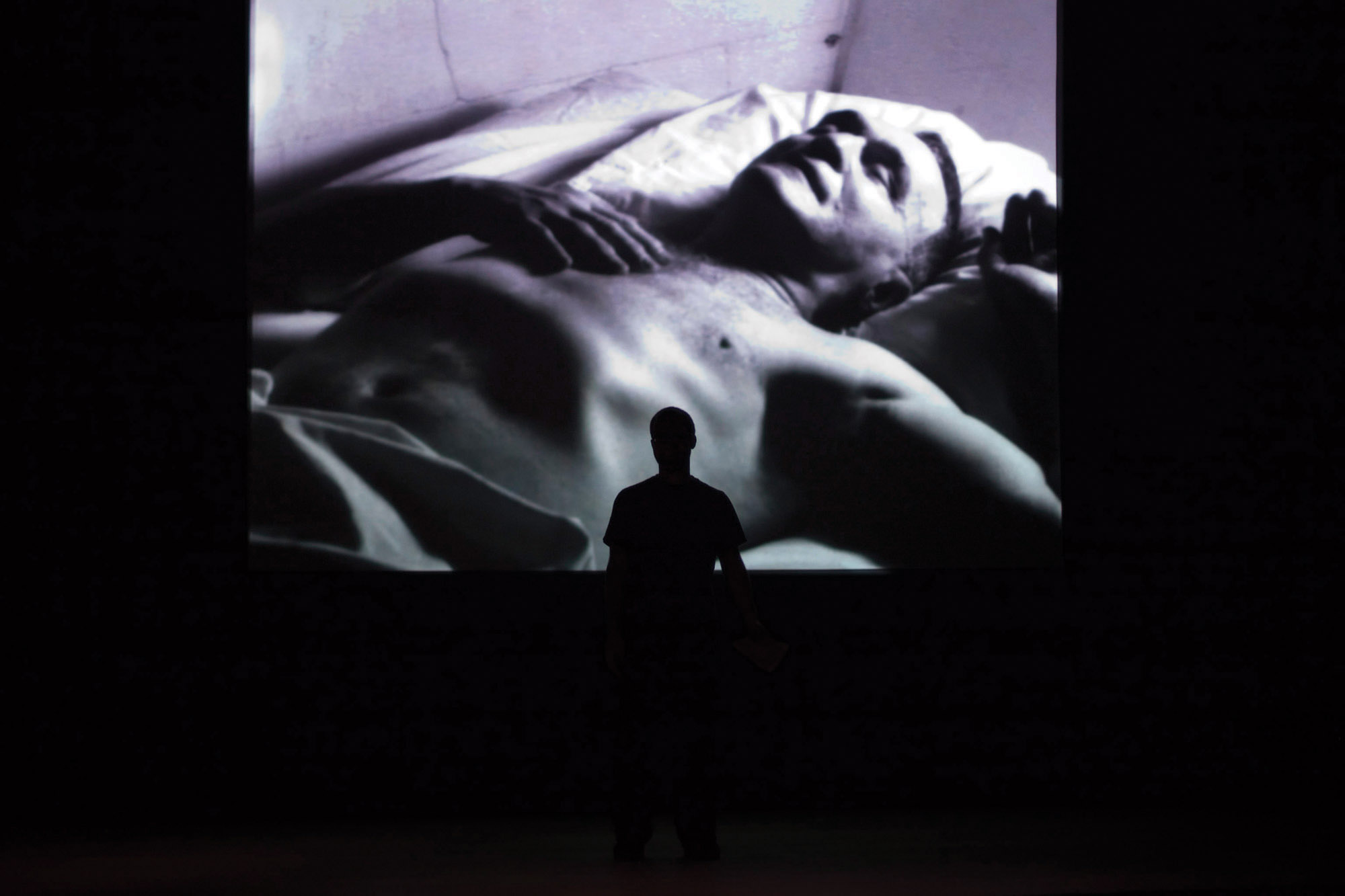 A man silhouetted against a large projection of a shirtless man sleeping. 