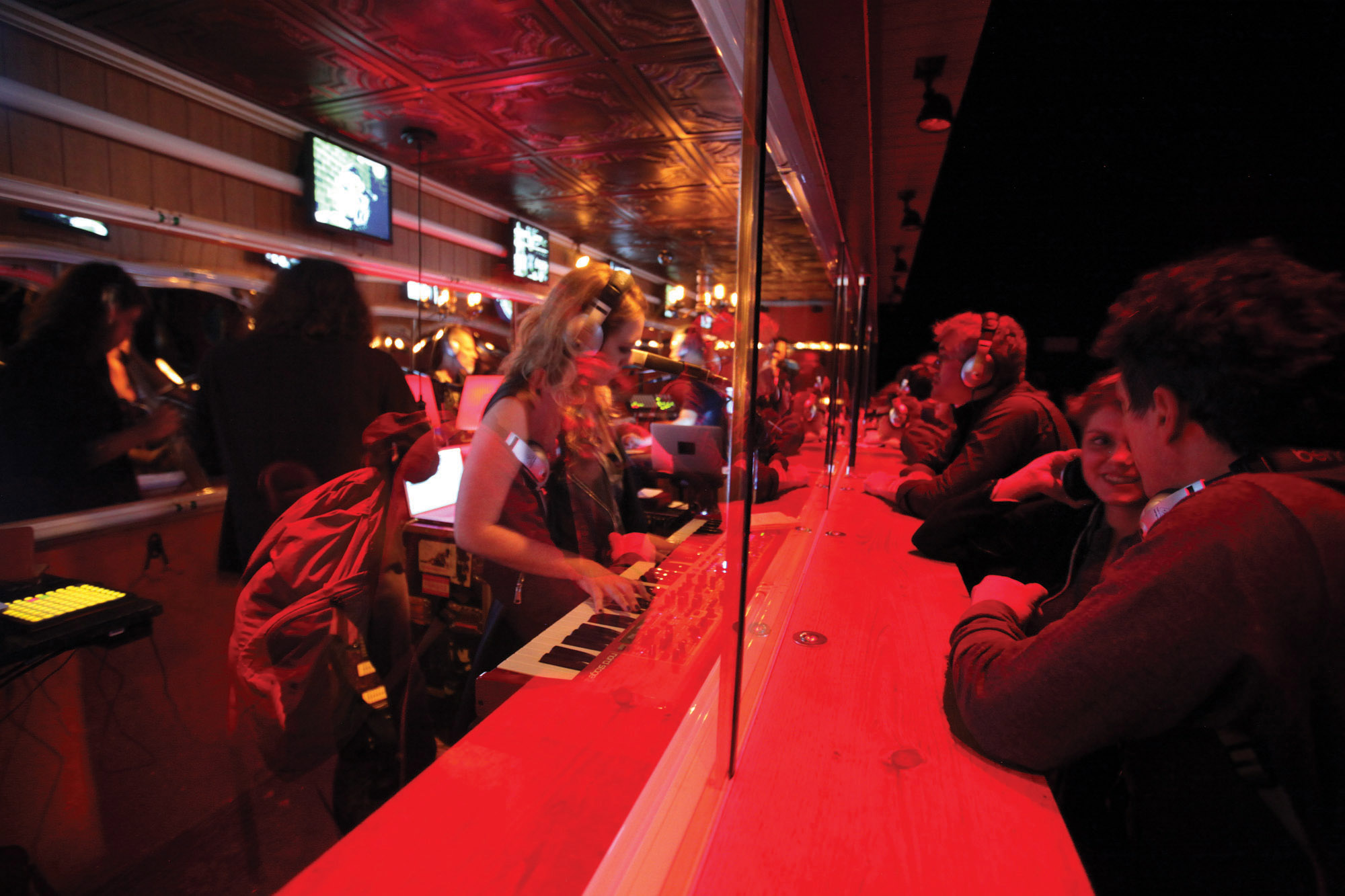 People sitting at a bar washed in red light separated by a pane of glass. On one side a woman is playing a keyboard and on the other folks are mingling and wearing headphones. 