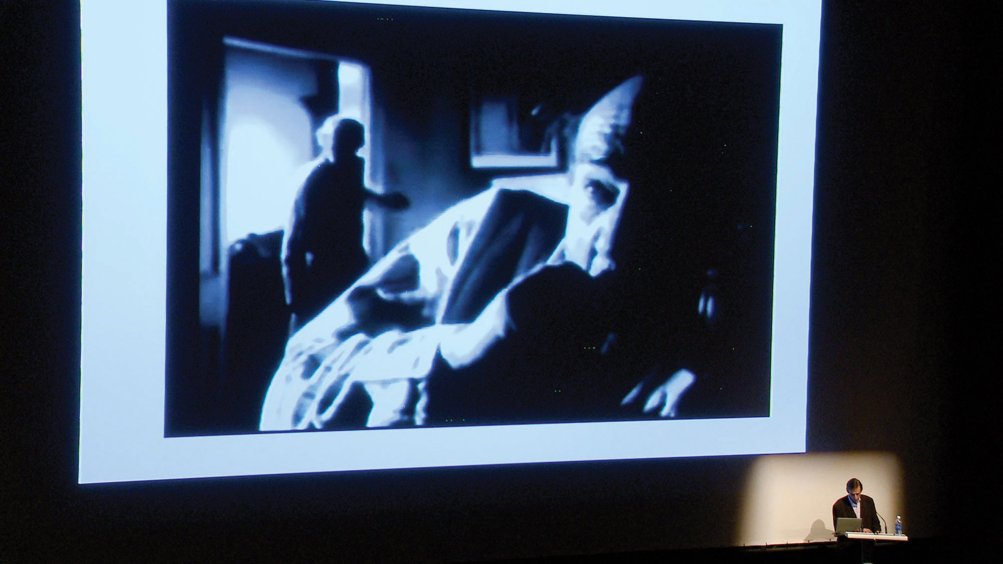 Steven Conner in fort of a large projection of a man, startled as another person enters the room. 