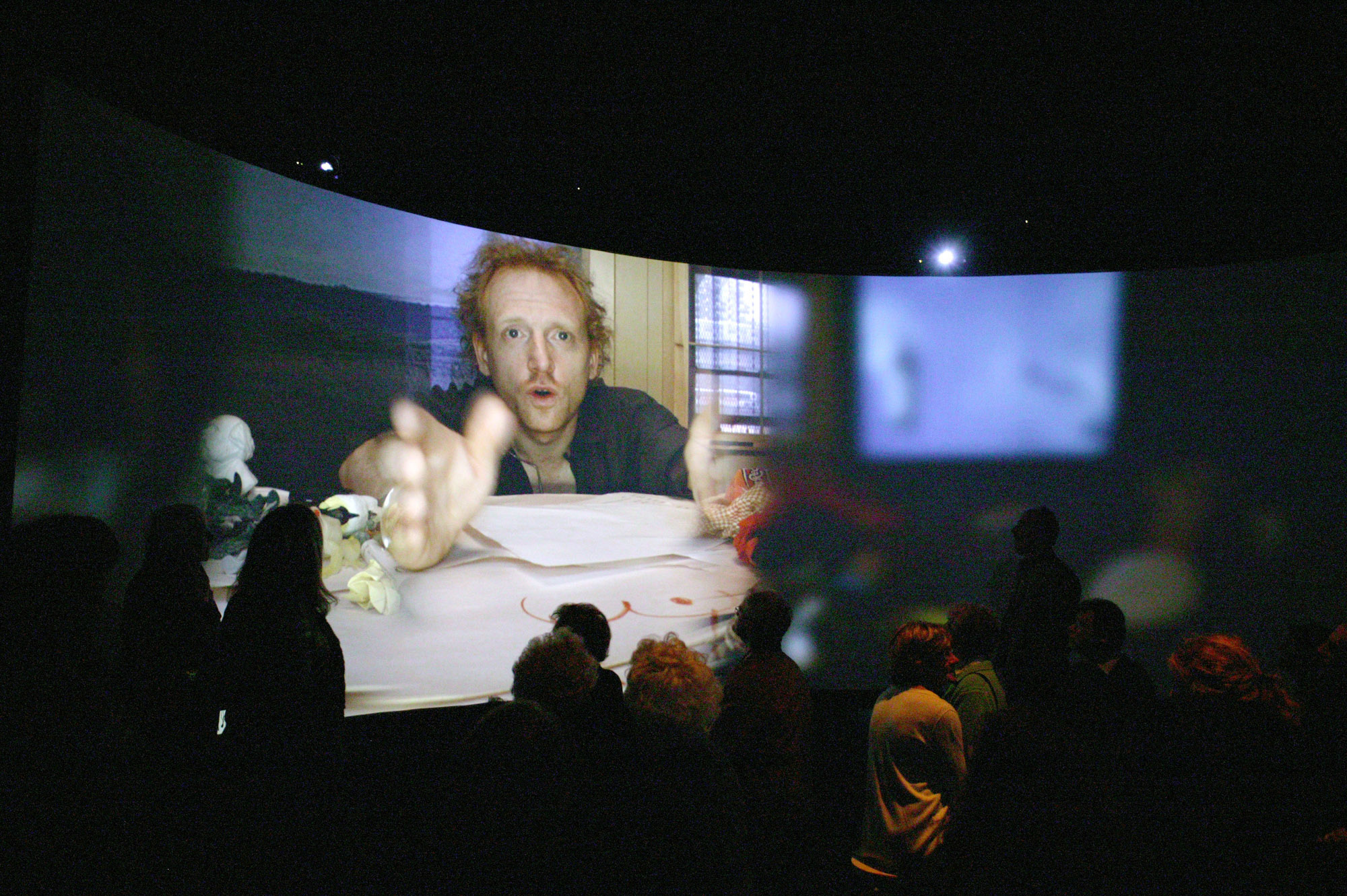 A small crowd milling about in front of a panoramic screen showing an image of a man with red hair reaching. 