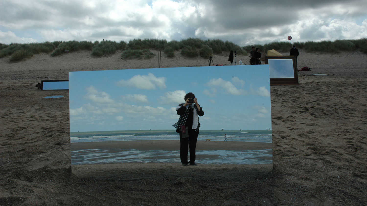 Agnes Varda taking a picture of herself in a large mirror placed on a beach. 