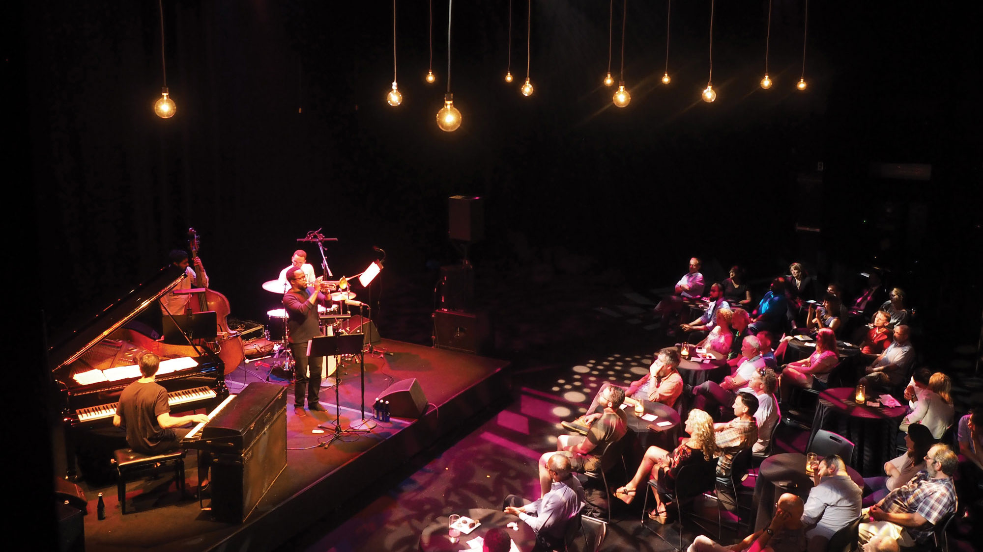 A jazz quartet playing on a small stage under a canopy round exposed bulb lighting infant of a small audience. 