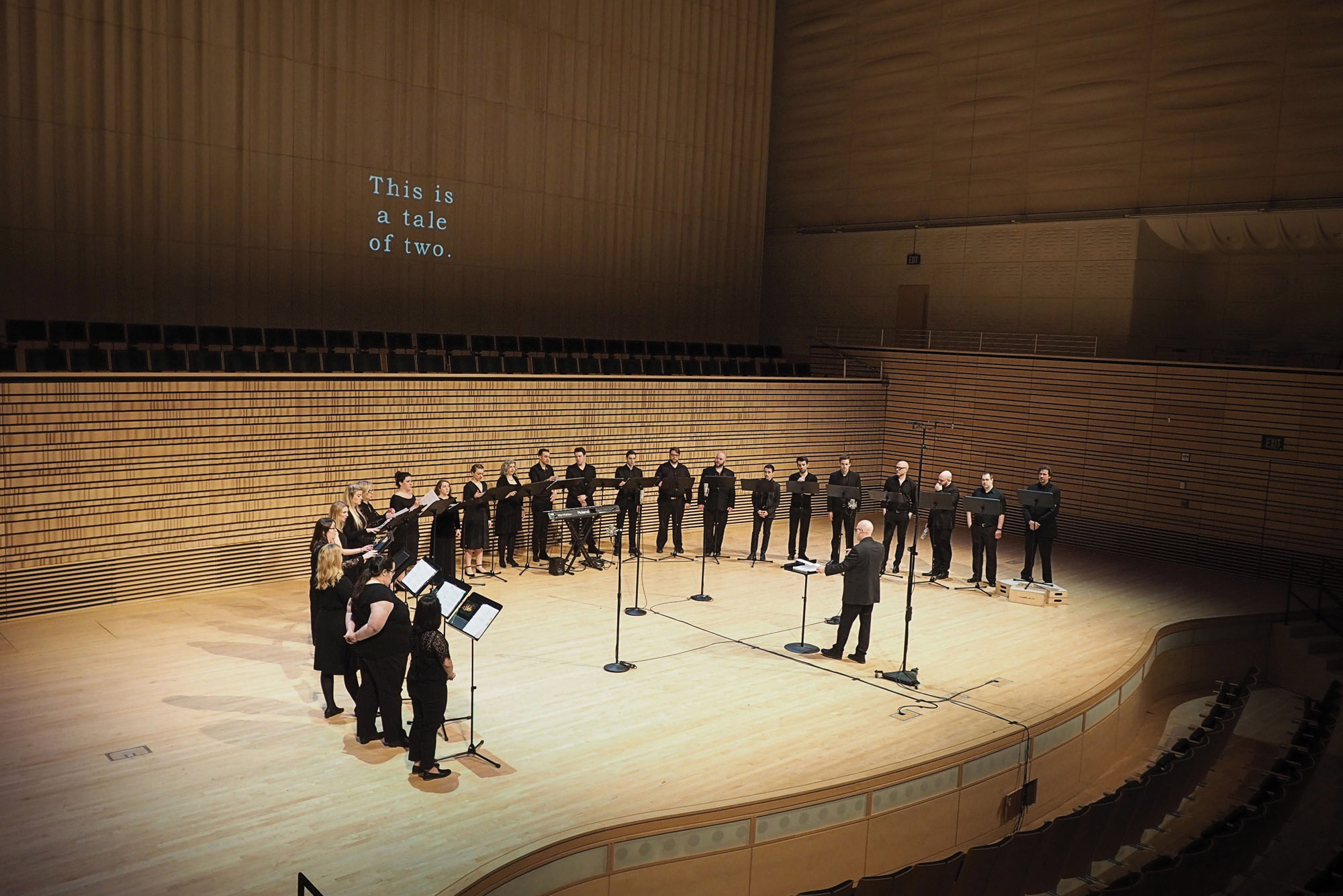 A choir dressed in black standing in a semi circle in front of a projection that reads "This is a Tale of Two" on the concert hall stage. 