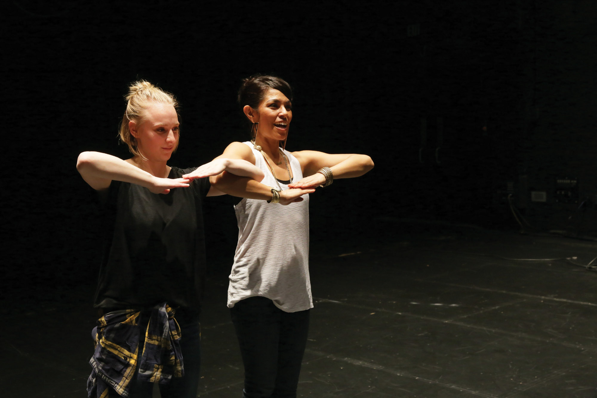 two women hold their arms in a bent T shape in a black box studio. 