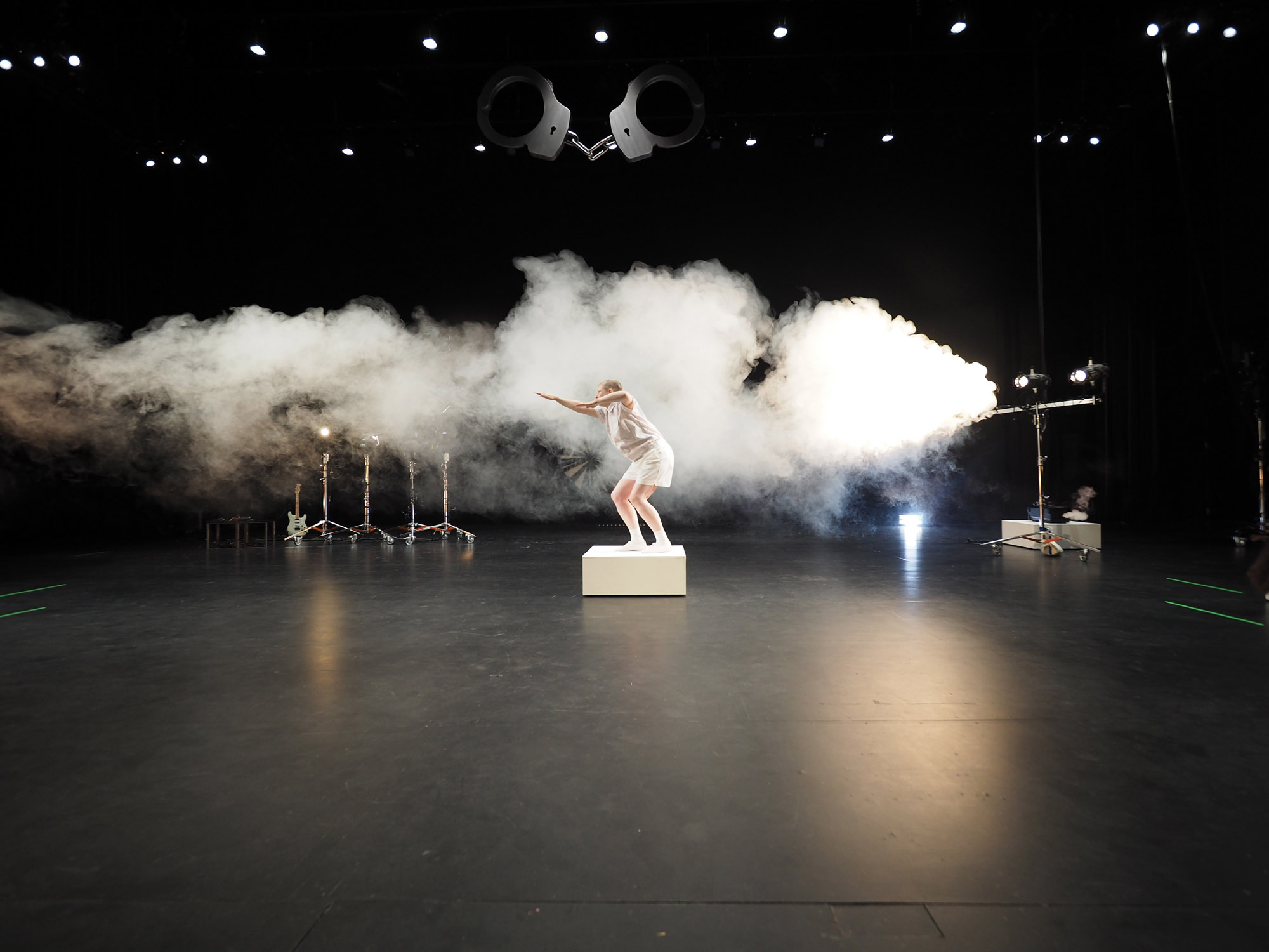 A dancer dressed in white standing on a white rectangular cube in front of white theatrical fog in a black box studio. 