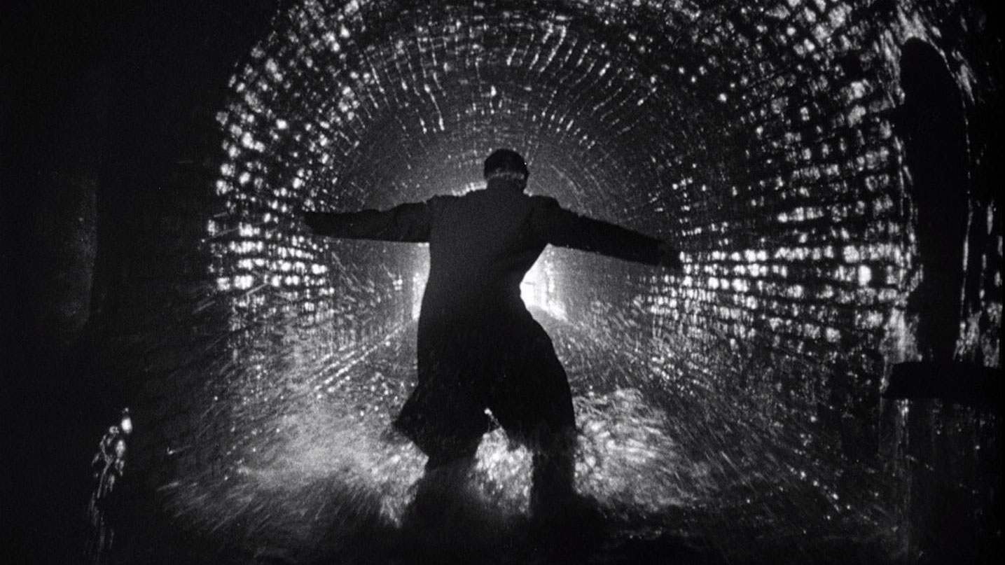 A man wearing a long coat with arms outstretched, touching each side of a cobblestone tunnel. 