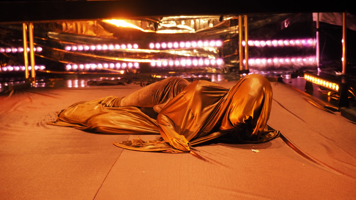 An unseen man in a shroud of brown silky fabric rolling on the floor in a studio lit pink lights. 