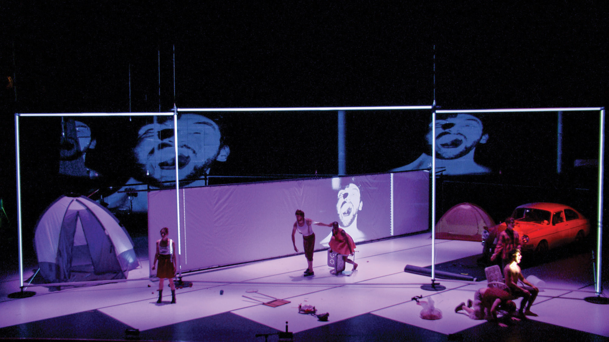 Six performers on a light purple stage with a vintage car, several tents, and other found objects.  Three performers are the front of the stage, one screaming theatrically. Two performers are behind them walking hunched towards the sixth performer.