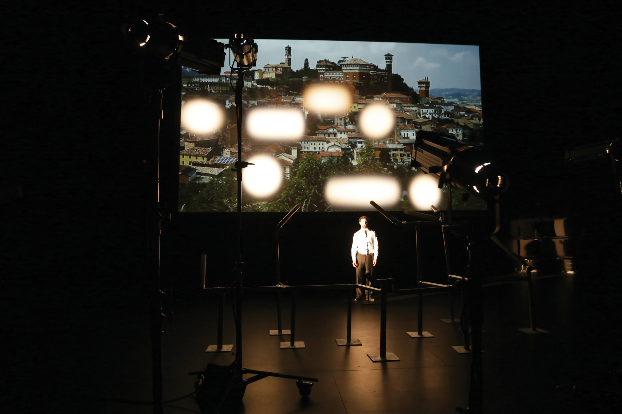 A man wearing a white button up shirt and black pants in front of various lighting rigs and a projection of a town's skyline. 