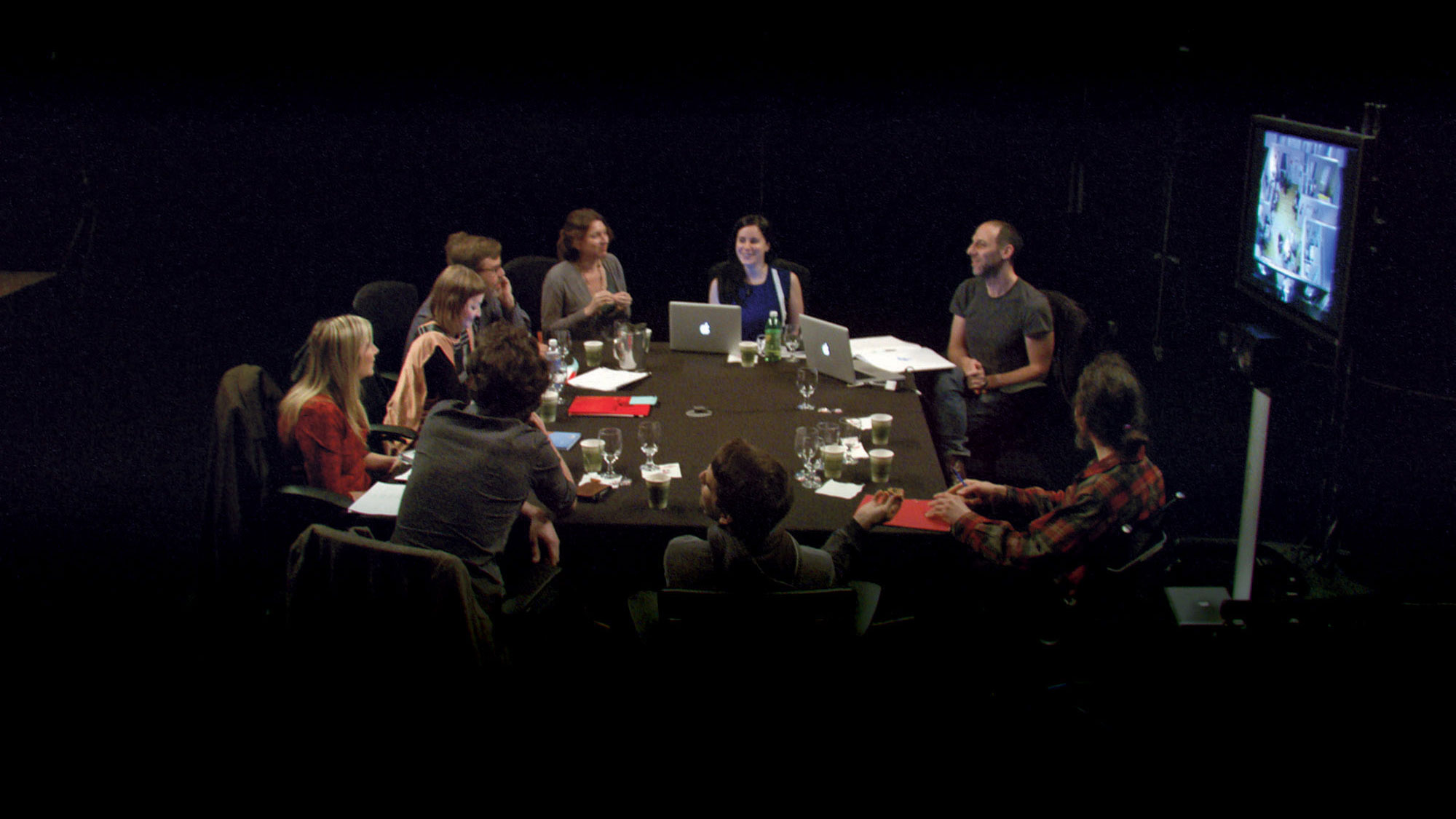 A group of nine people sitting around a conference table in active discussion in a dark room. 