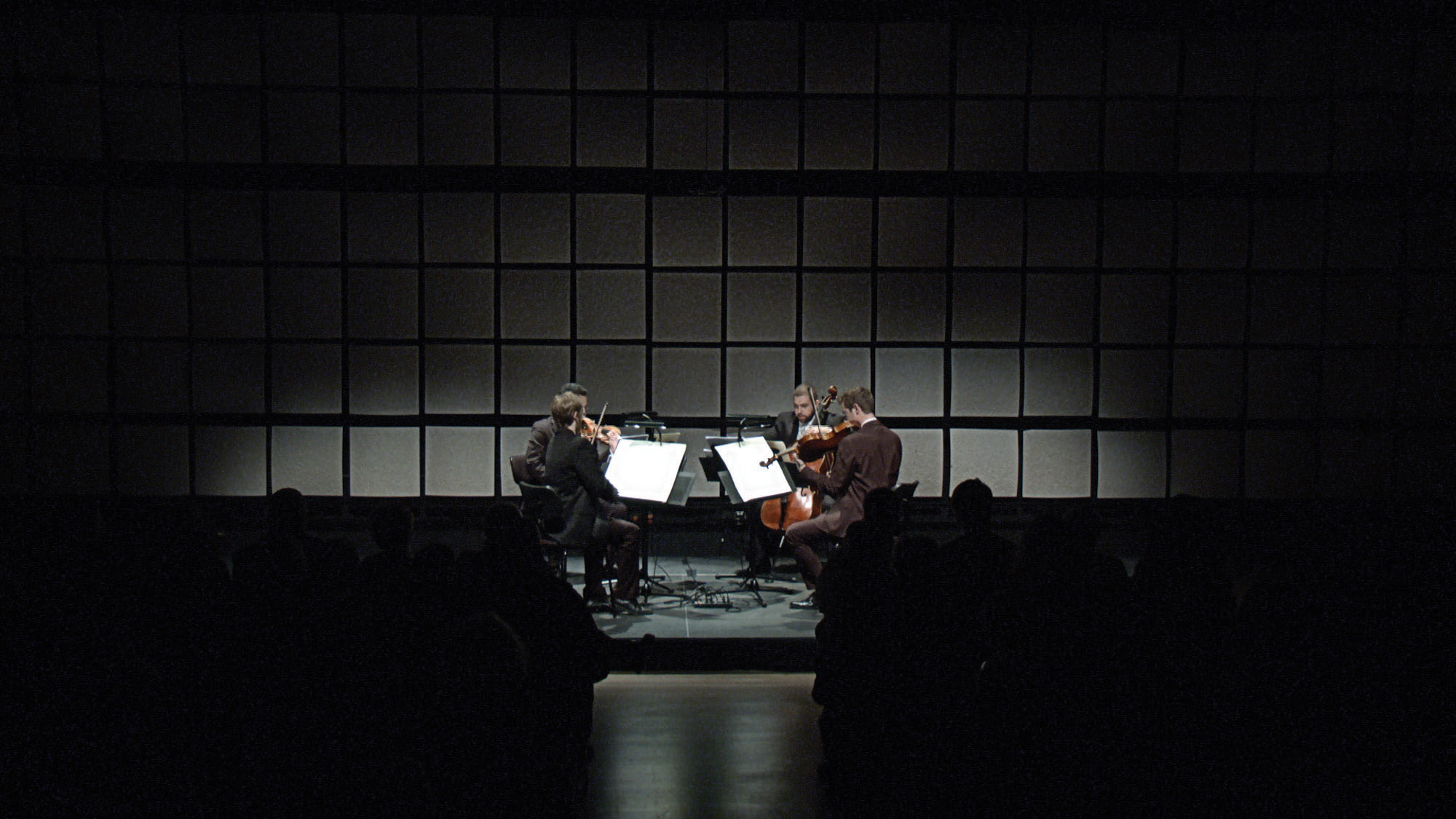 A quartet in a right formation performing a concert on a dark stage in front of a wall of gray acoustic tiles. 