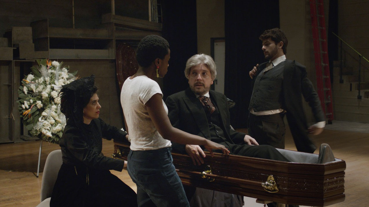 Two people in Victorian era mourning clothing and one woman in modern clothing gathered around a coffin. A man with white hair is sitting up in the coffin talking to the people around him. 