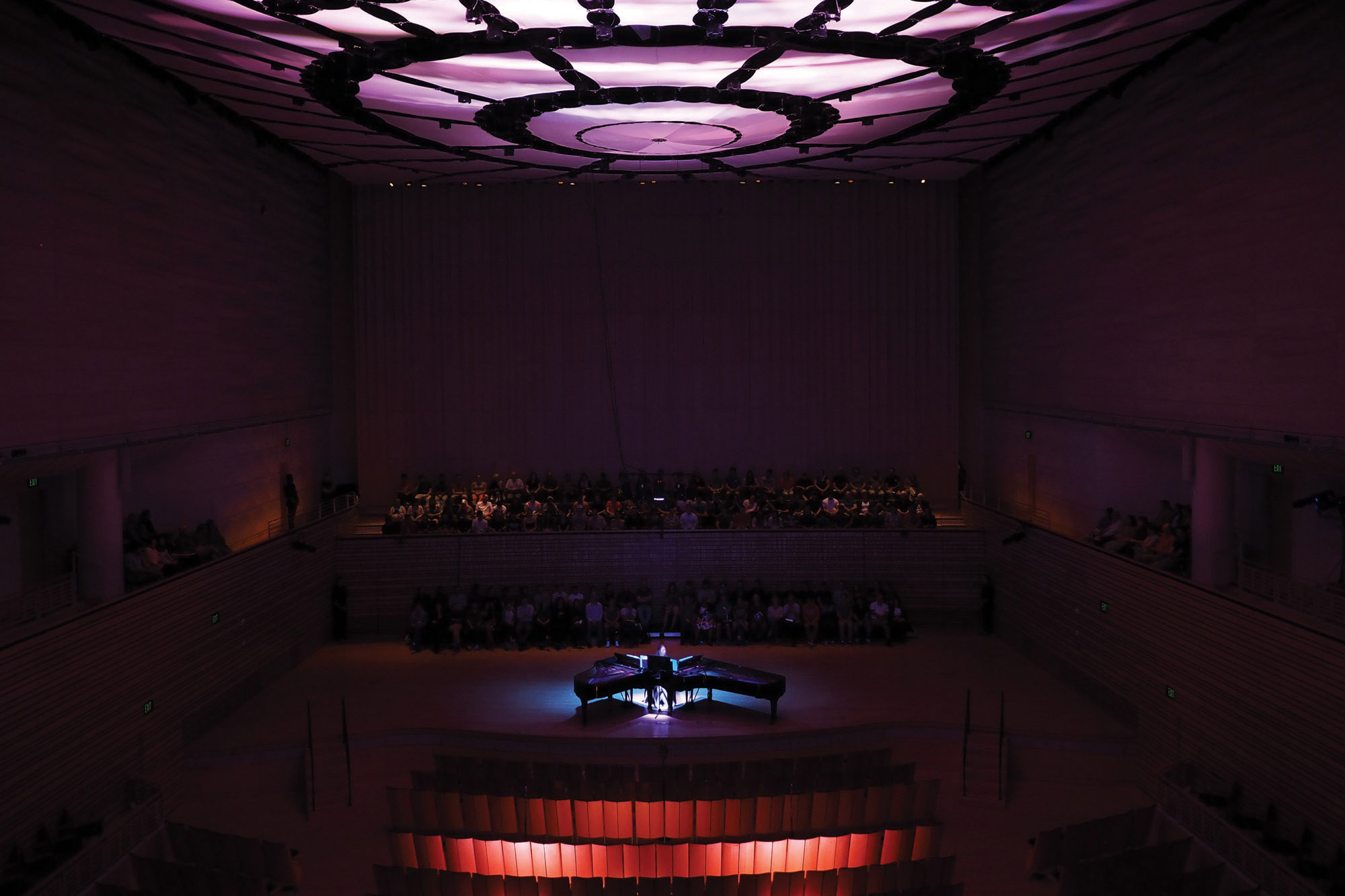 A person playing two pianos on the concert hall stage washed in purple and red light with a crowd behind them. 