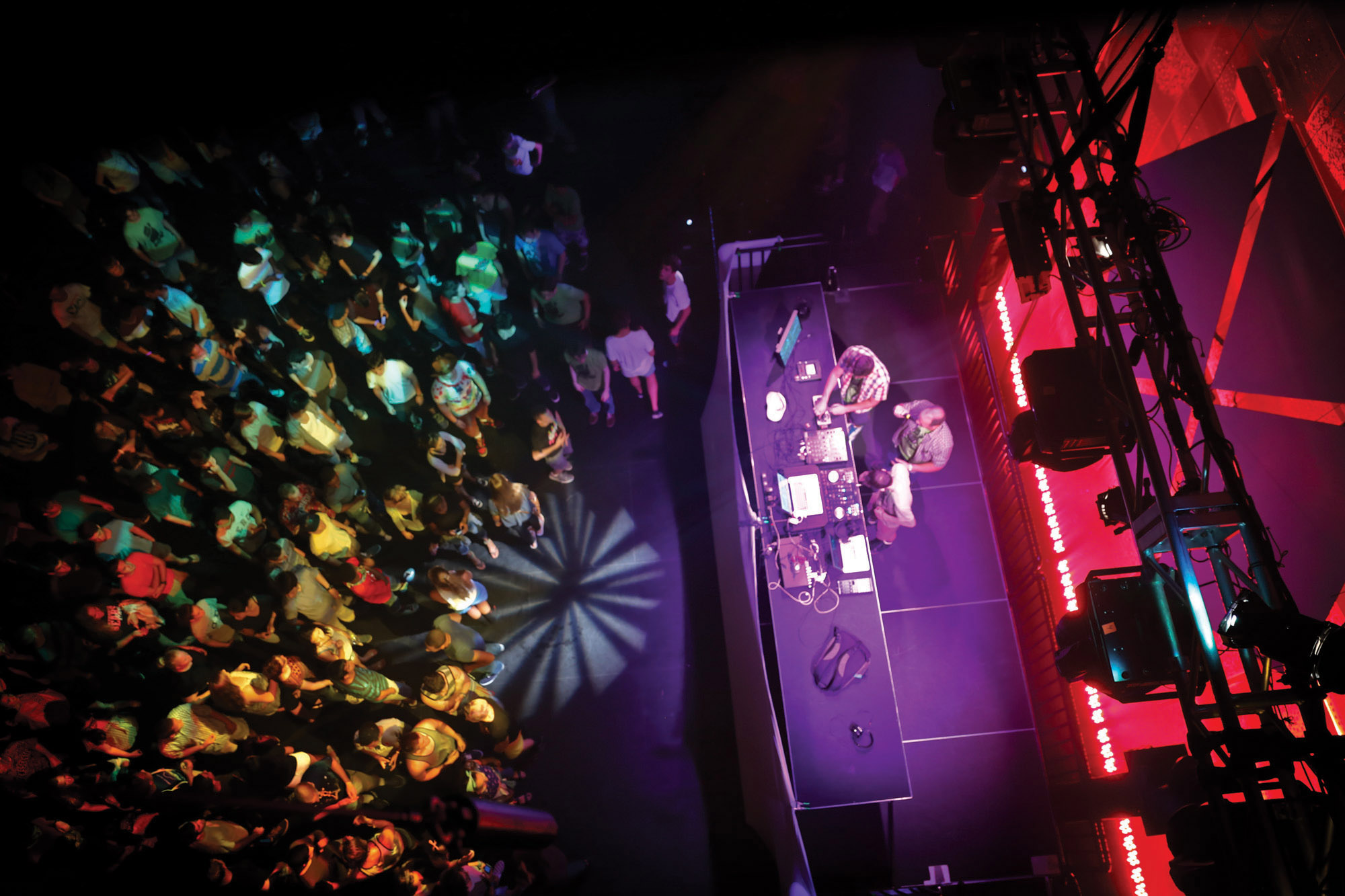 An aerial shot of three men DJing on stage performing for a crowd in a wash of rainbow party lights. 