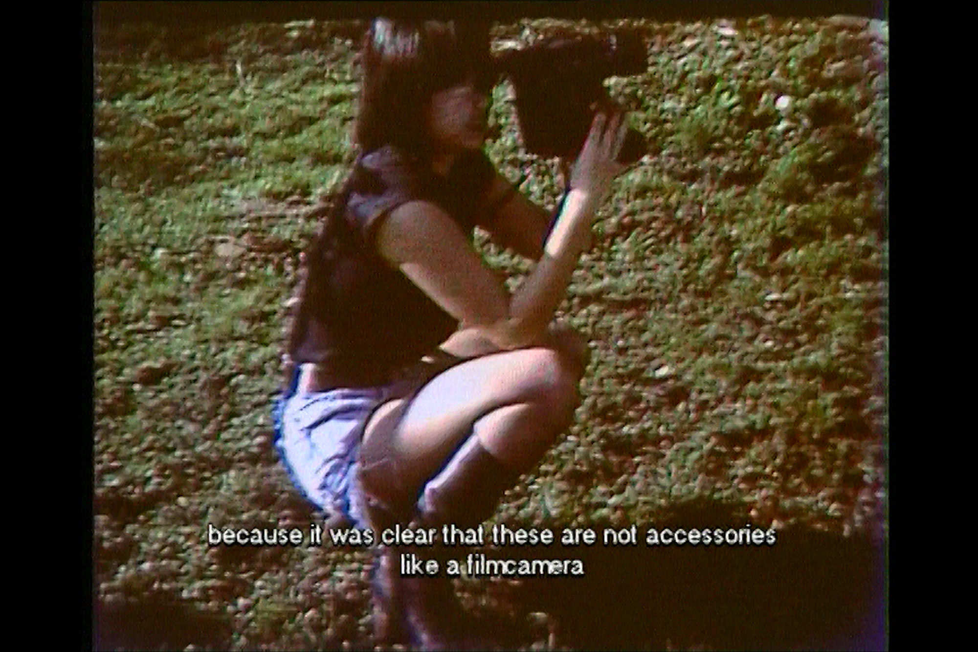 An asian woman wearing a black top and jean shorts squatting in the grass shooting with a VHS camera, Because it was clear that these are not accessories like a film camera. 