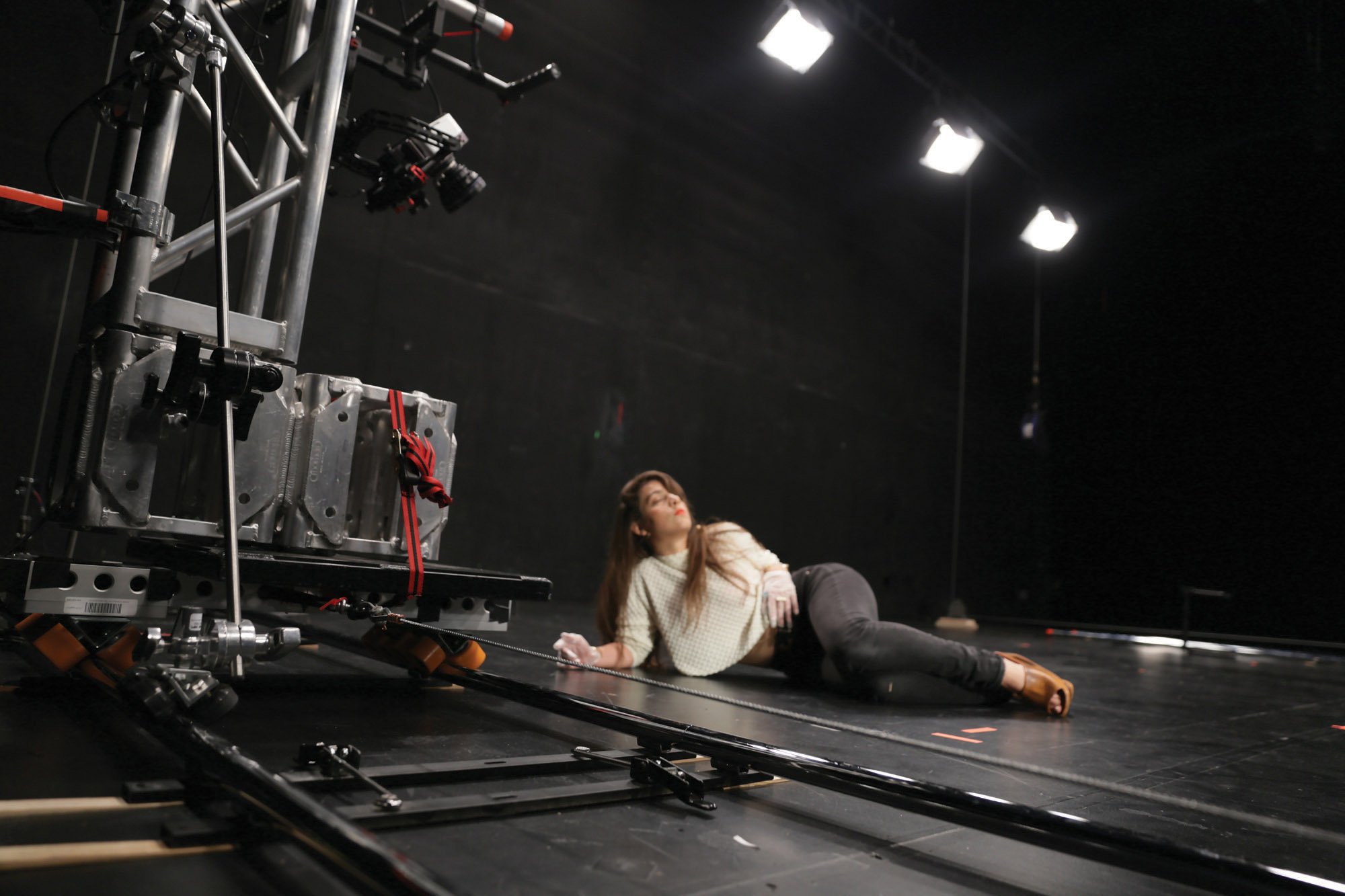 a woman with brown hair wearing a cream sweater and black pants laying on her side on a black stage. 