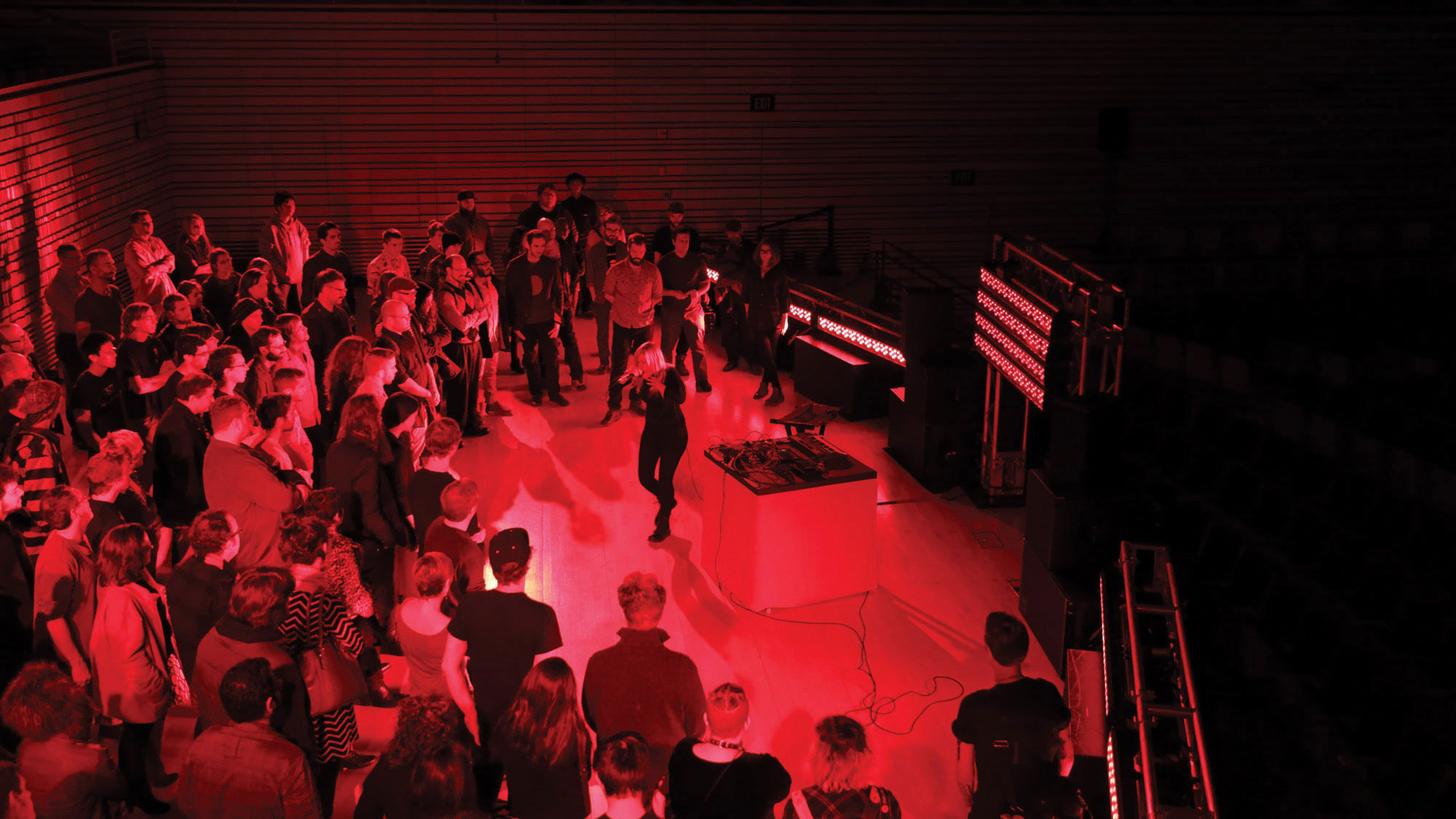 Pharmakon performing on the concert hall stage with an audience standing around her washed in red light. 