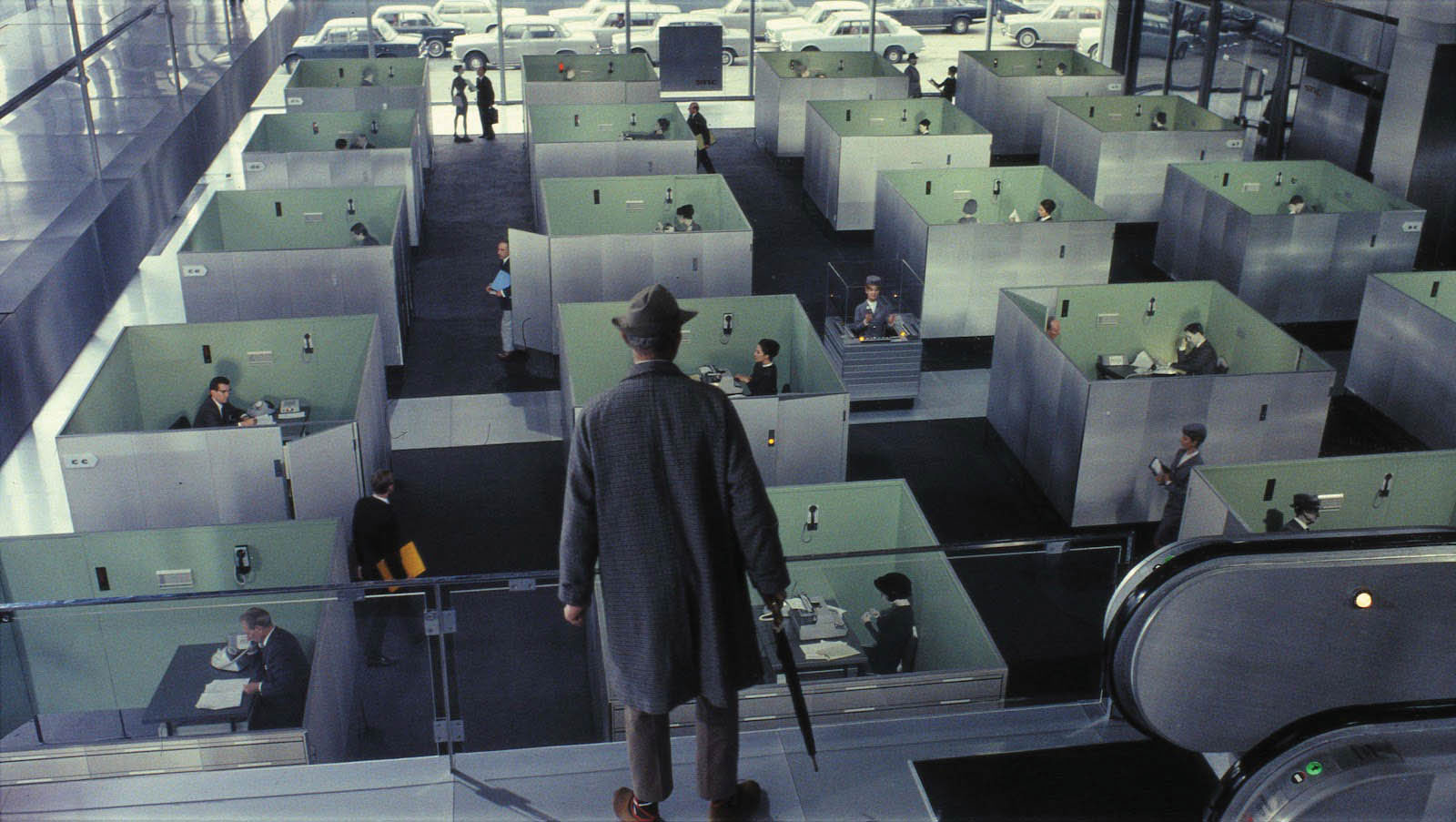A man wearing a 60's era trench coat and fedora standing above an office of cubicles looking down at the workers. 