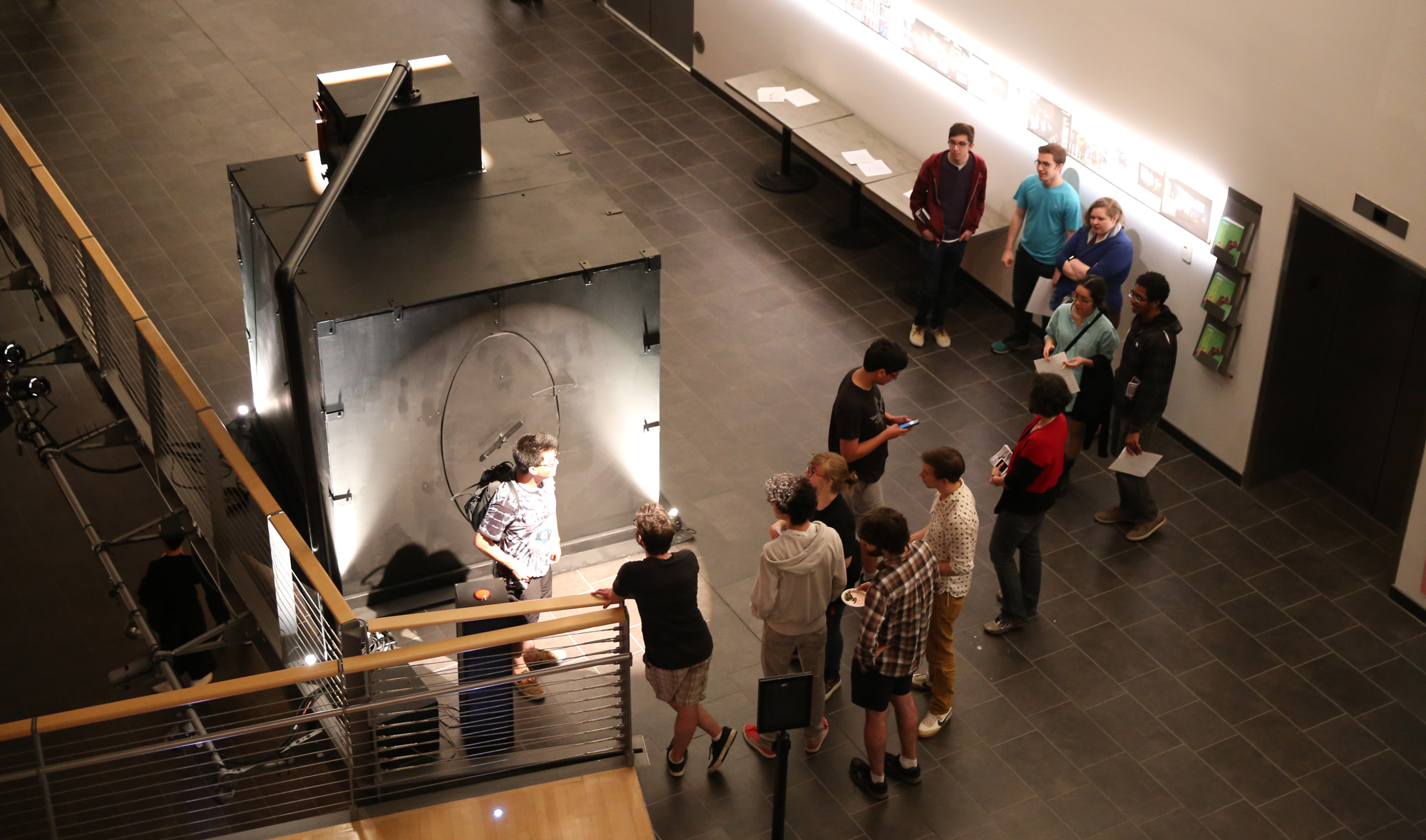 Students waiting in line out side of a black box with an oval door in a hallway. 