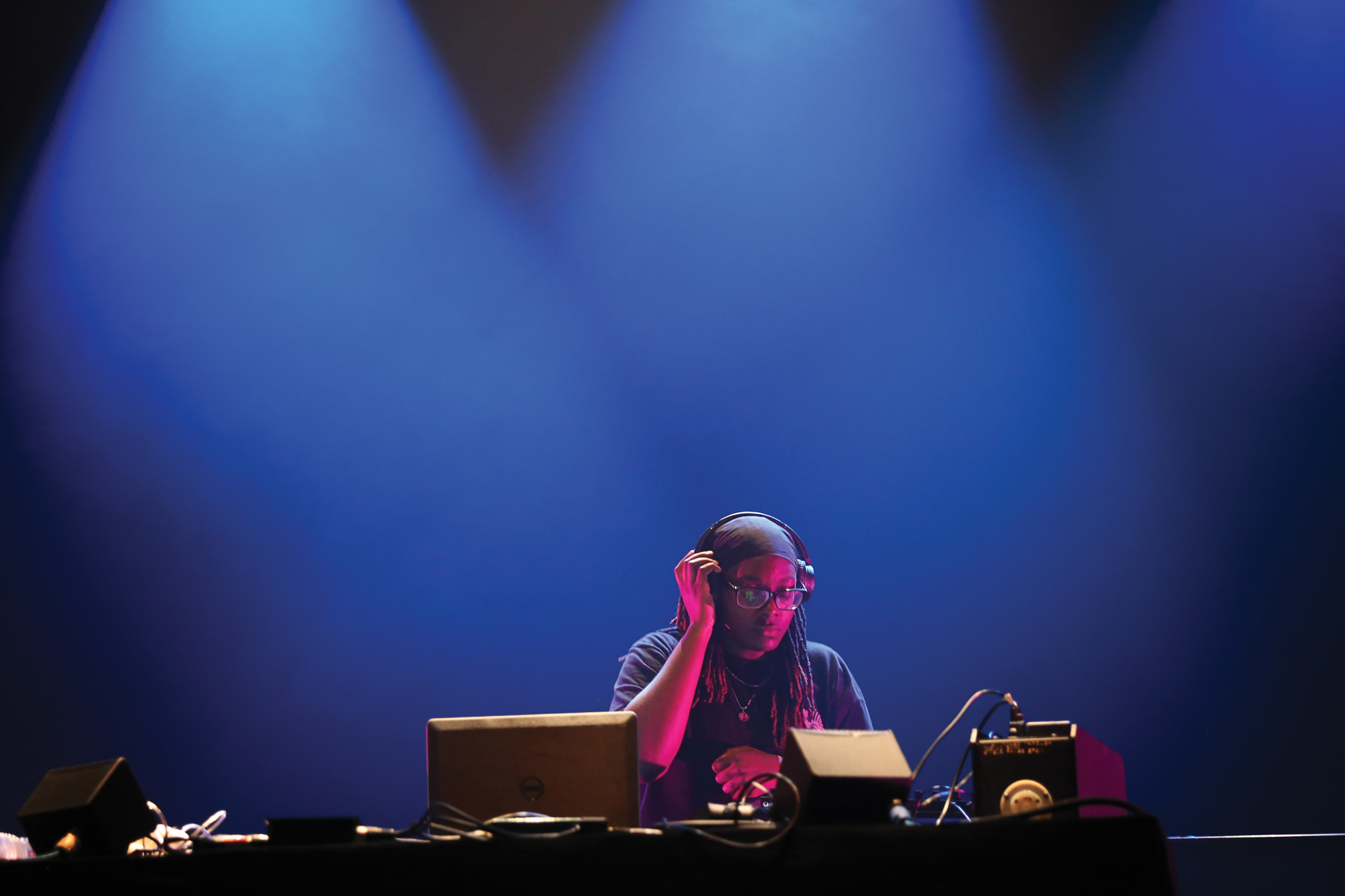 A Black person wearing glasses with their hand on one ear of their headphones, DJing in front of a wall of blue light. 