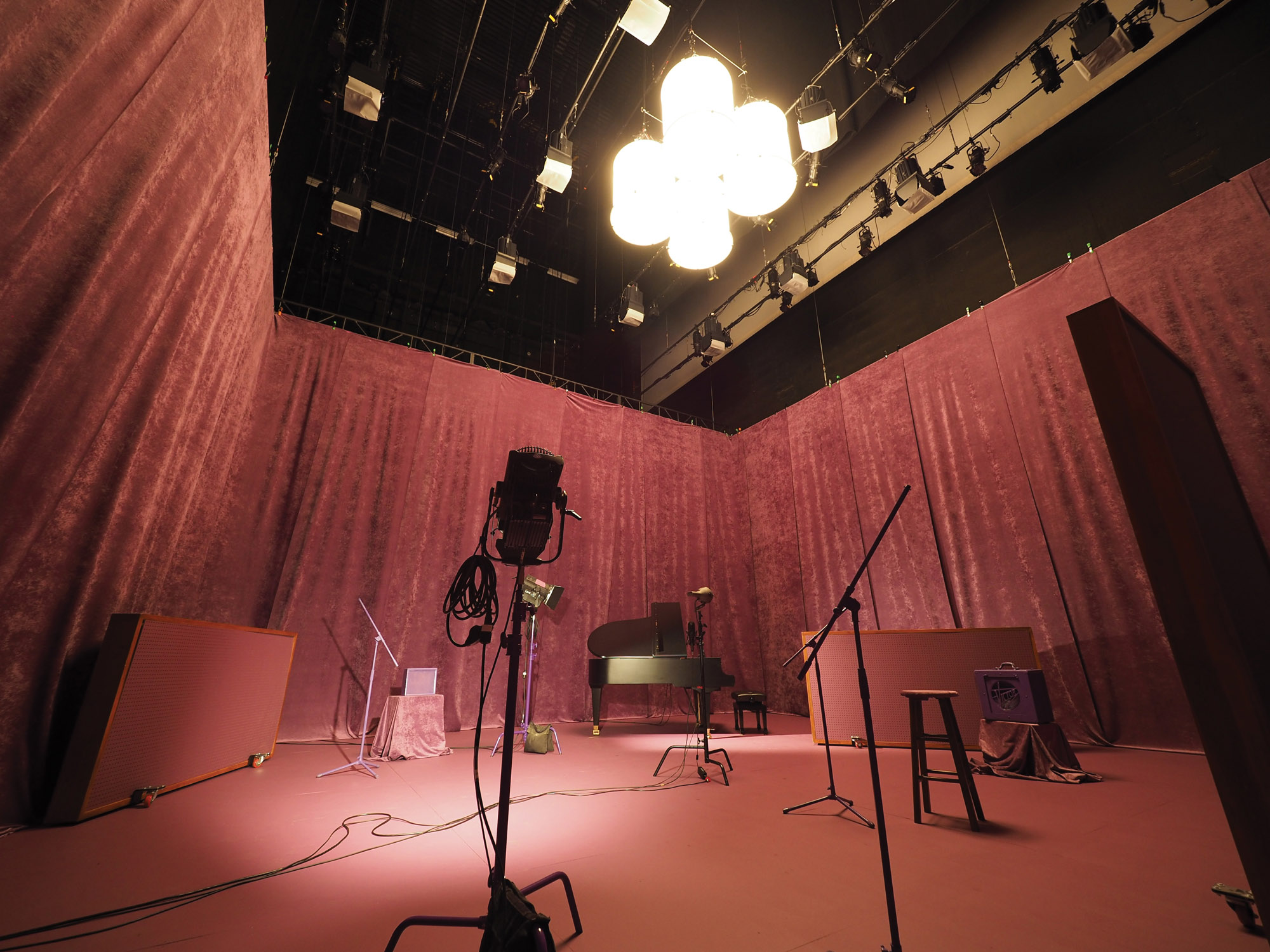 Studio set up with a piano, microphones, amp, and various lighting in a room with pink velvet walls with matching pink floors