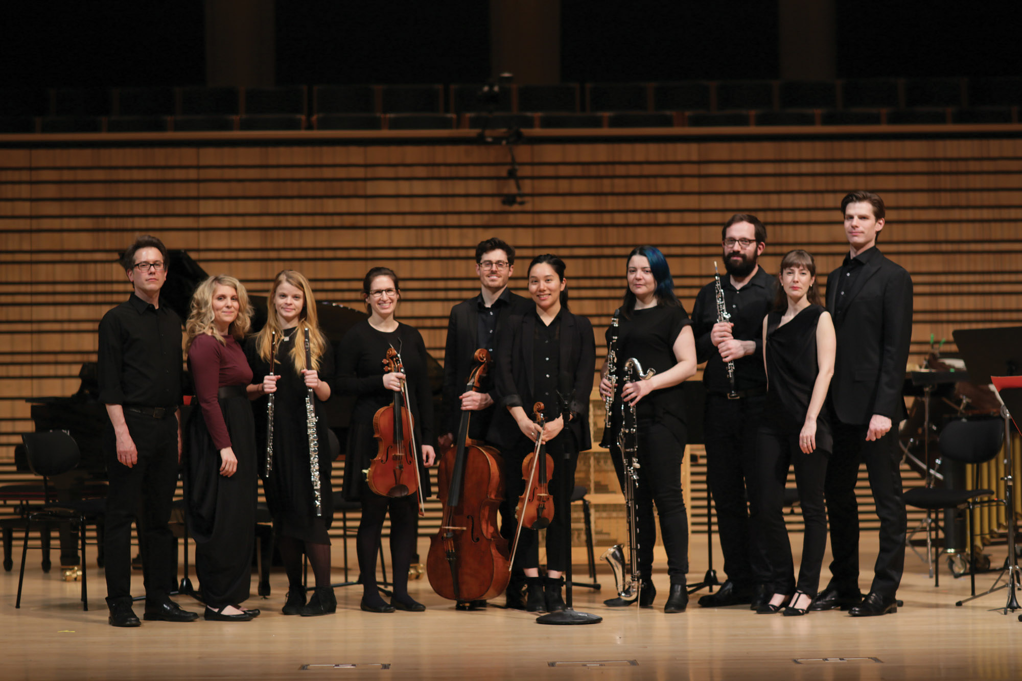 A small orchestra dressed in black posing for a picture, smiling at the end of the concert hall stage. 