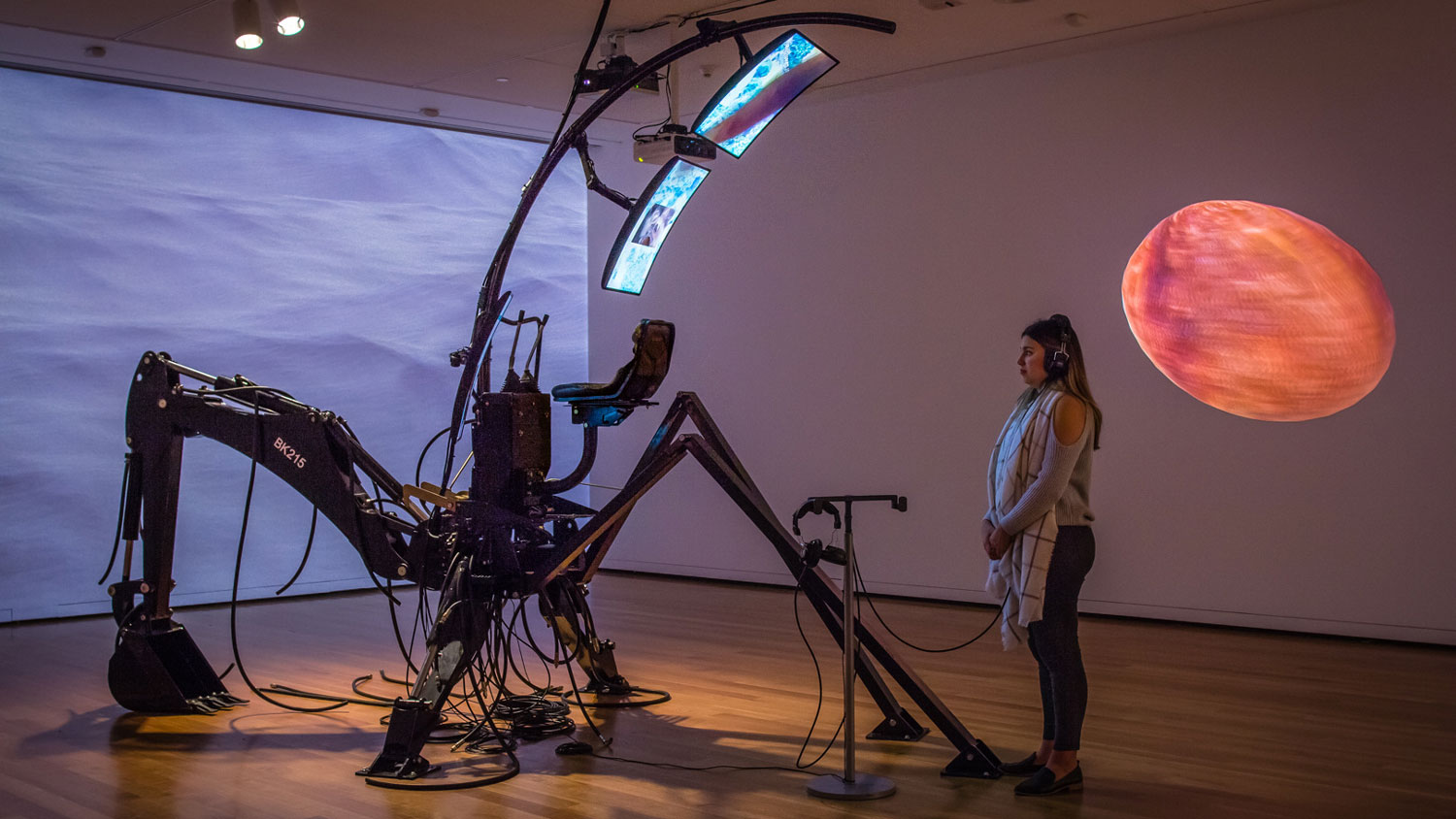 A woman wearing headphones looking at two screens held by a spider-like apparatus or sculpture in blank room with a projection of the ocean and a planet. 