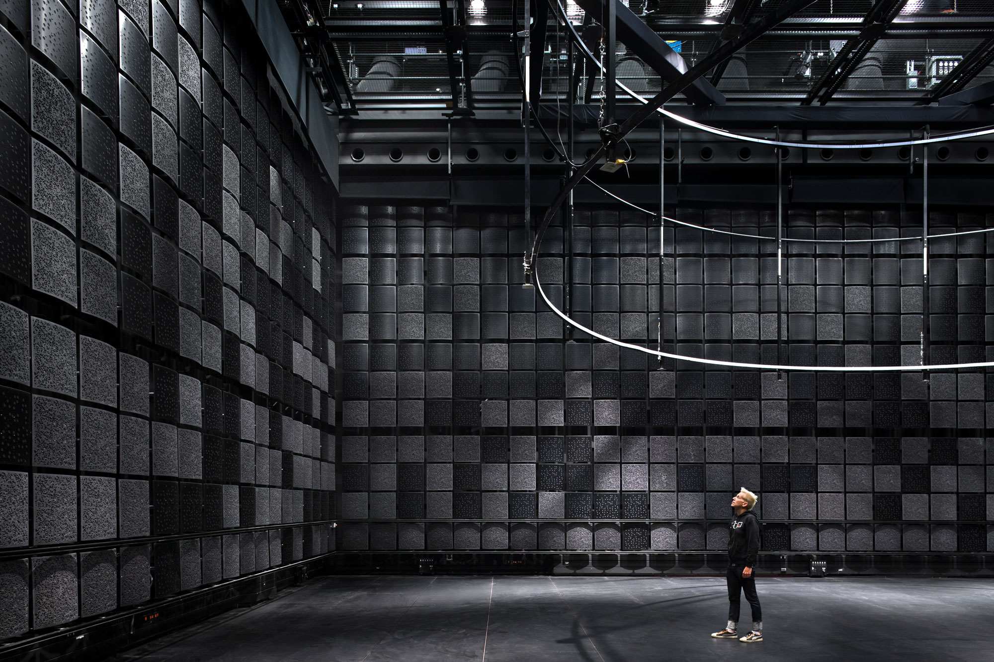 A white person standing in a black box studio looking at a wall of black sound proofing squares. 