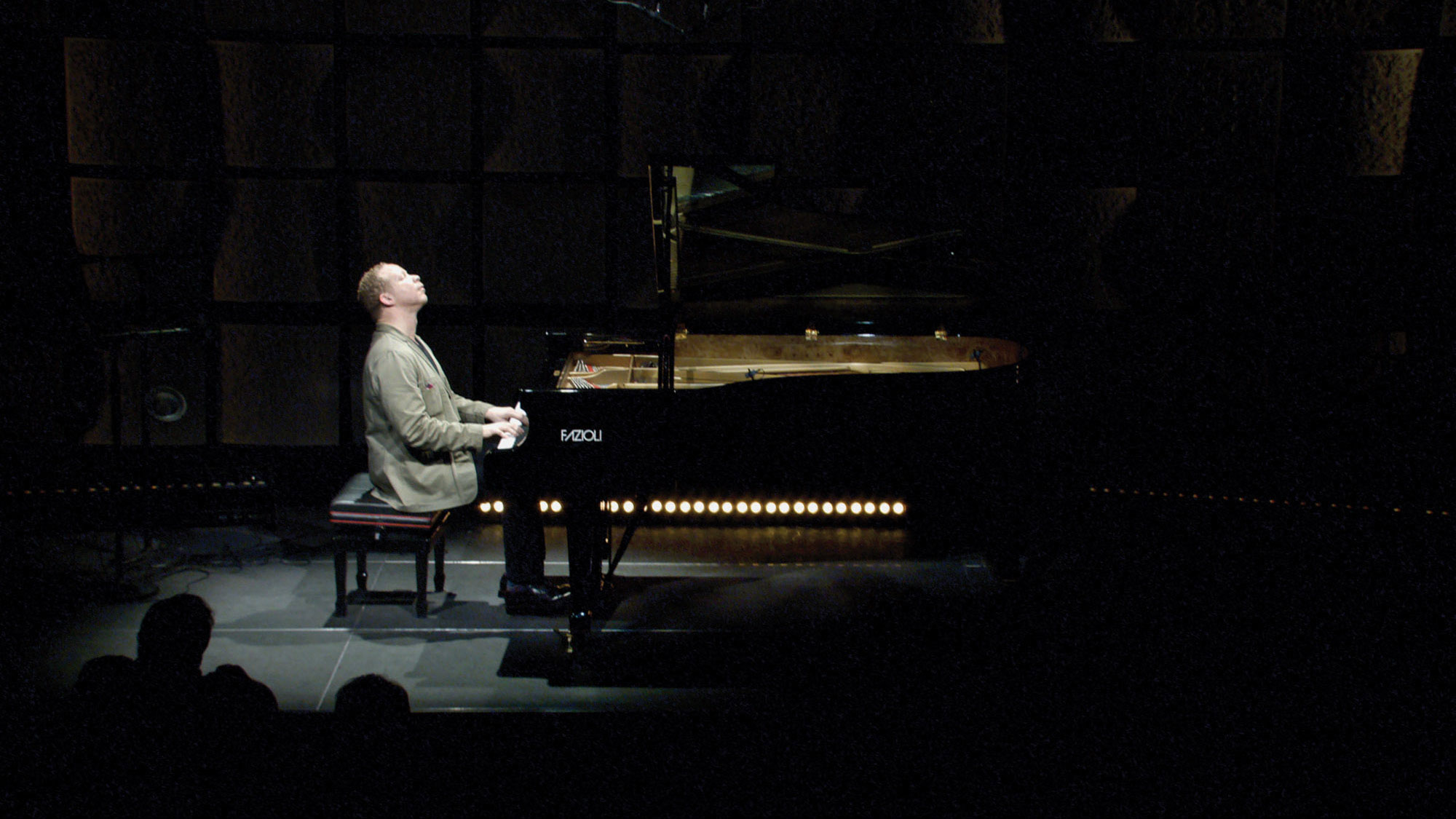 A man lifting his head towards the ceiling with eyes closed in a moment of reverence as he plays a grand piano on a dark stage in a spotlight. 