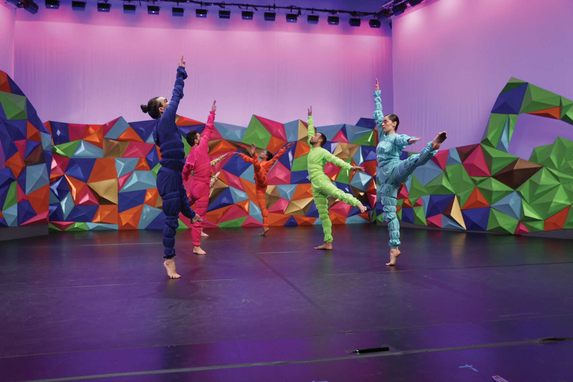 Five dancers in purple, pink, orange, lime green, teal outfits dancing in front of colorful geometric wall 