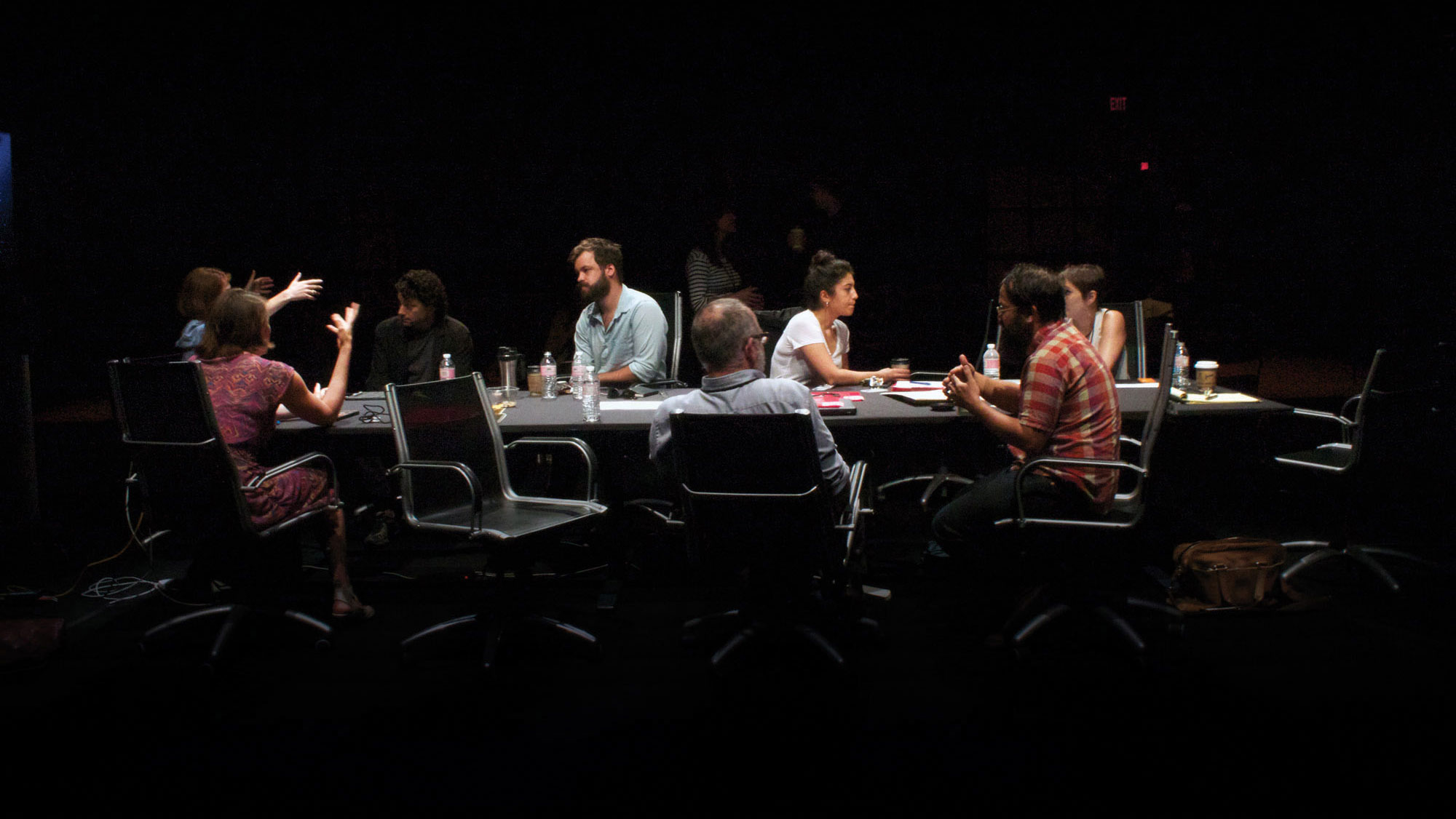 A group of eight in active discussion sitting around a conference table in a darkened room. 
