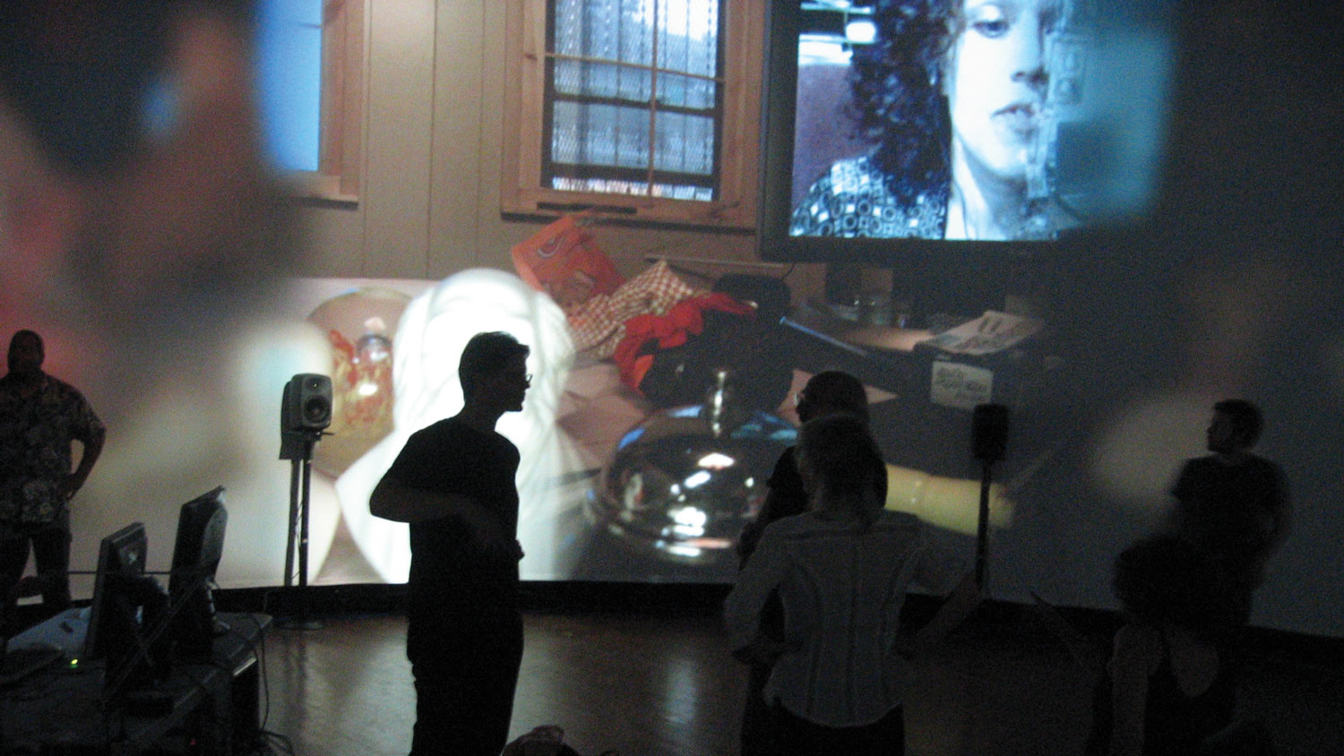 Three people chatting intently silhouetted against a large projection of a cluttered room. 