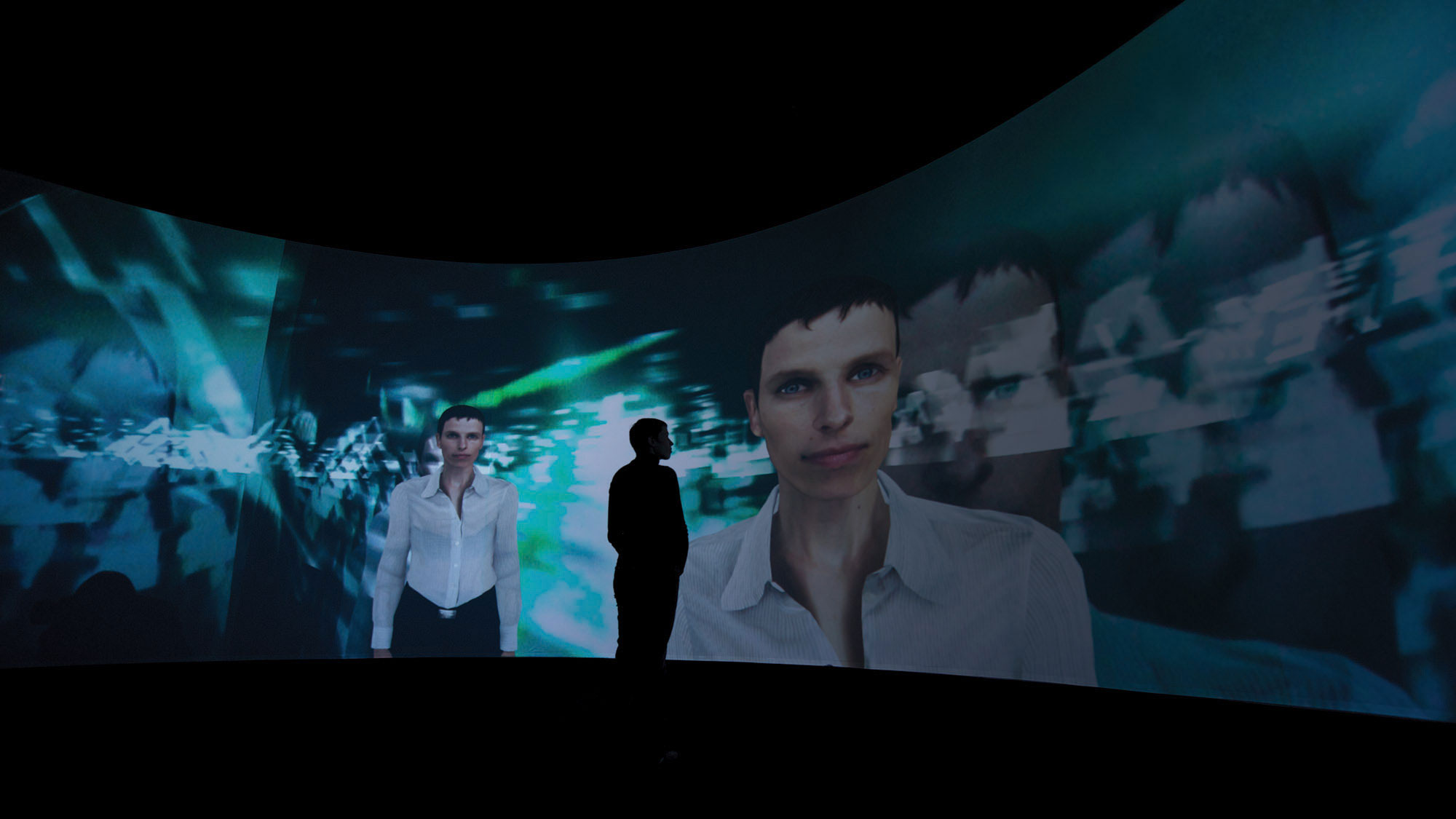 A computer generated image of a woman with a pixie cute and wearing a white button down shirt projected on a panoramic screen as a silhouetted person looks on. 