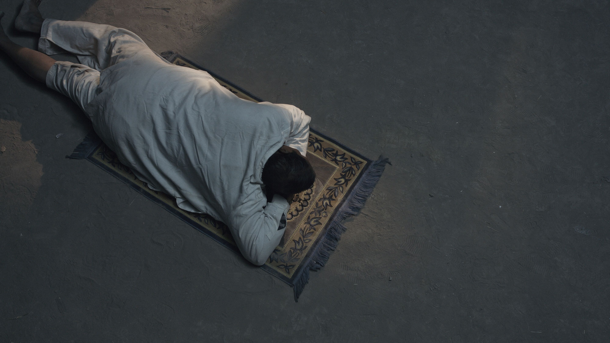 A man wearing draped white fabric laying in prostrate on a small prayer rug on a gray concrete floor. 