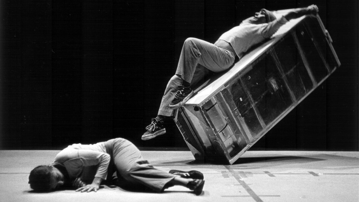 a female dancer lying on stage as a male dancer lays on a refrigerator that balances on it's corner 