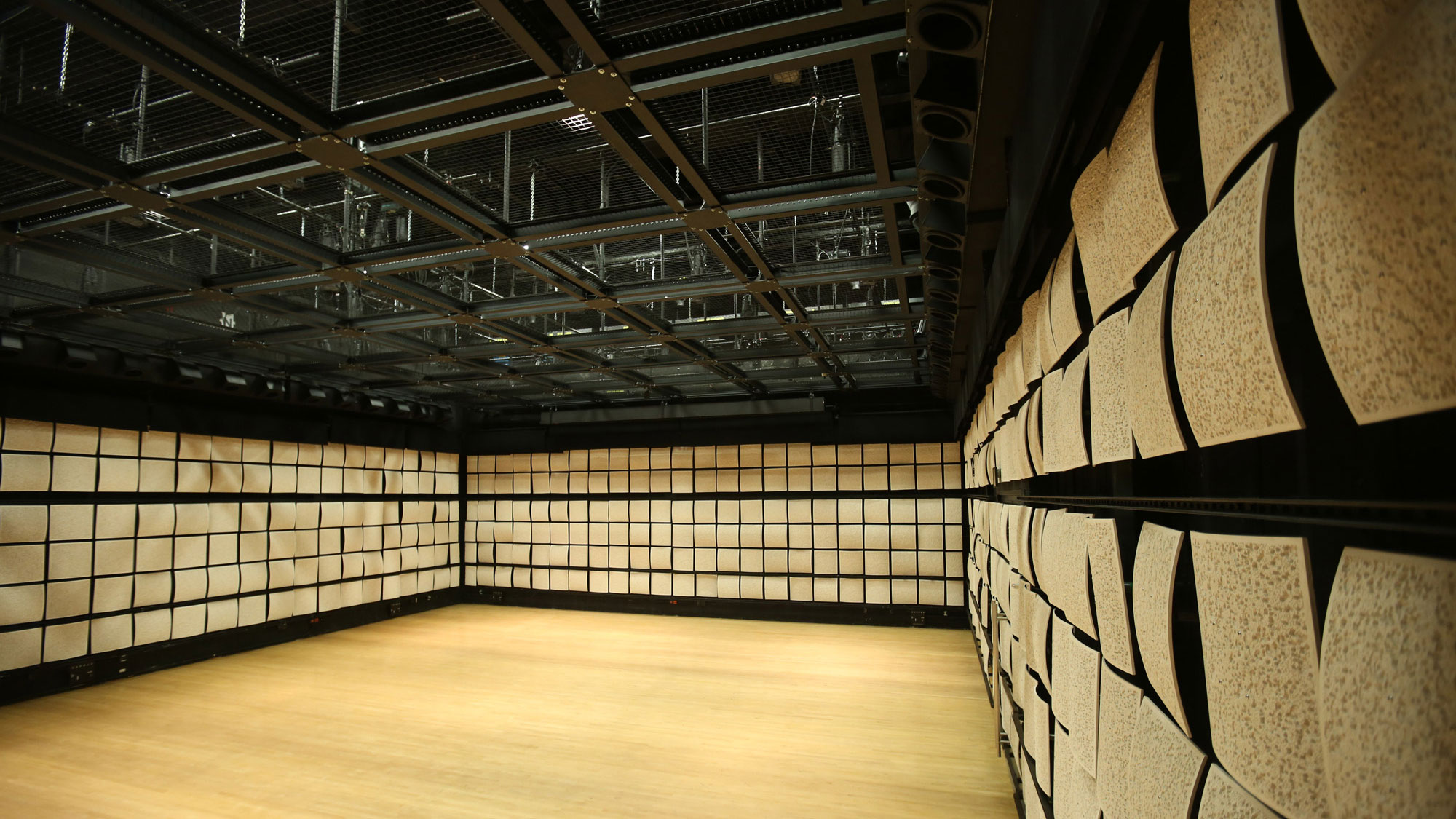 An empty studio with light wood floors, tan acoustic tiles on the walls, and black rigging on the ceiling. 