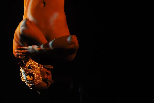 A shirtless Black man with arms crossed hanging upside down wearing a bedazzled gas mask. 