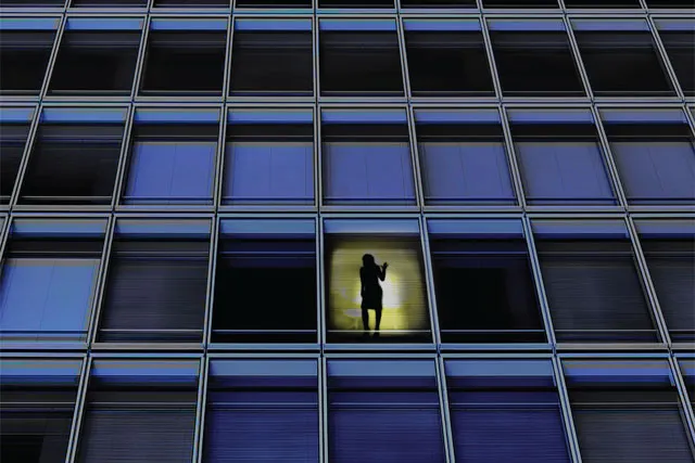 A high-rise building of blue tinted windows. A woman is silhouetted in a window lit by yellow light.  