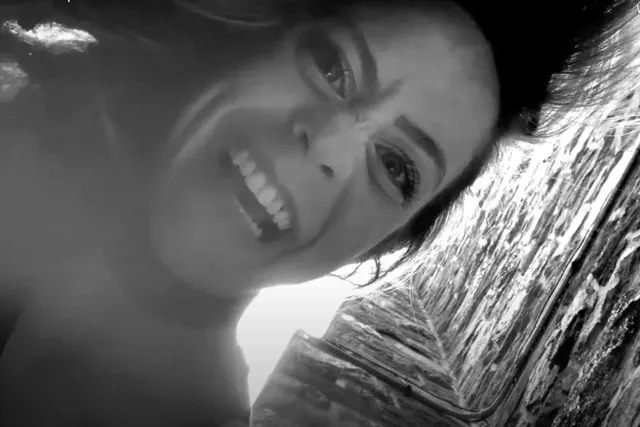 A white woman laughing manically down, into the camera while standing near a cobblestone wall.