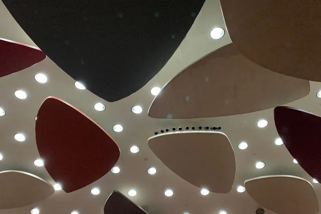 Rounded triangular geometric shapes in muted primary colors mounted on to a white ceiling with rounding lights in between. 