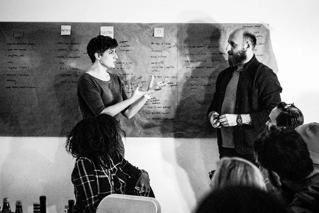 Annie Saunders talking to a bearded man in front of a blackboard in front of a small crowd. 