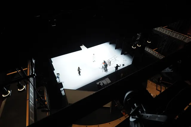 An aerial view of a dark theater with a white stage as a crew works behind the scenes. 