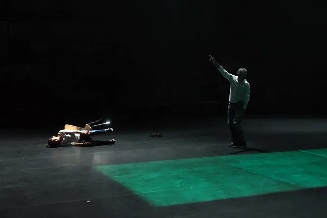 A man standing on a black stage pointing up as another person lays on the floor tipped over in their chair. A green rectangle is projected on to the floor. 
